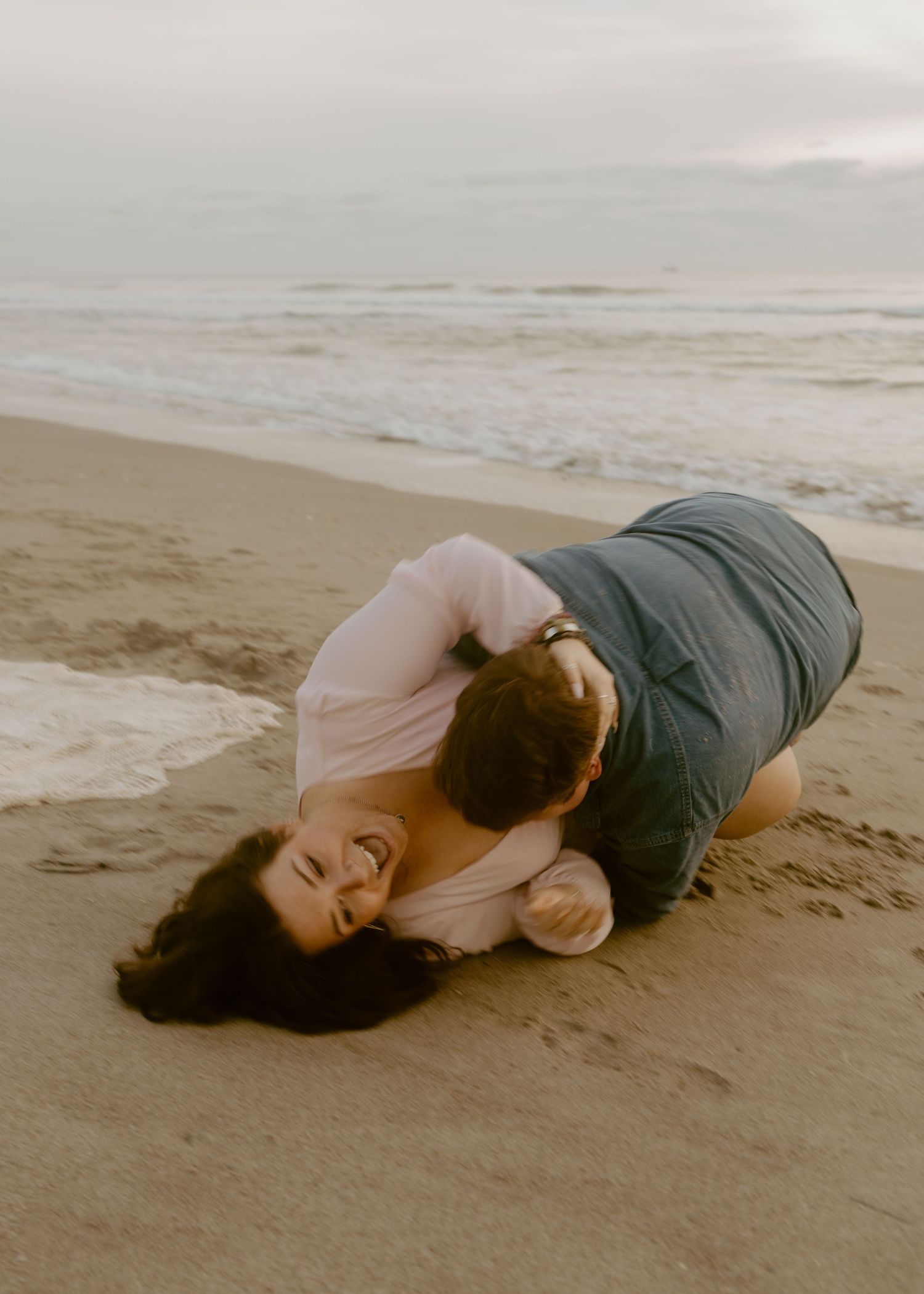 Couple laughing and cuddling on sandy beach shore, waves in background, romantic candid moment.