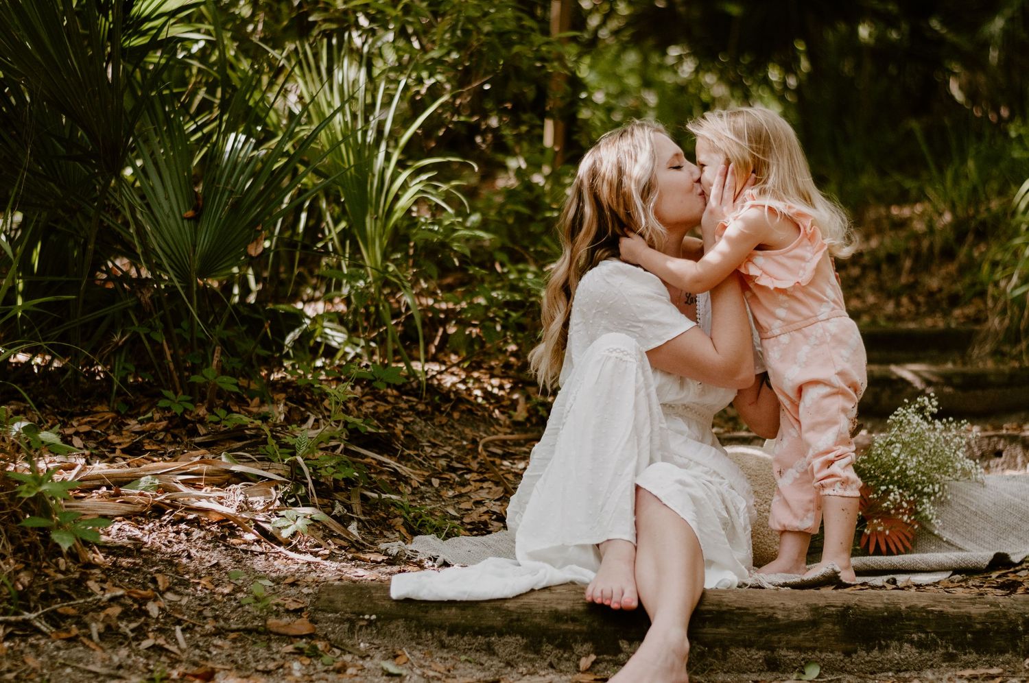 A mother kisses her young daughter outdoors in a lush green forest setting, both wearing white and pink outfits.