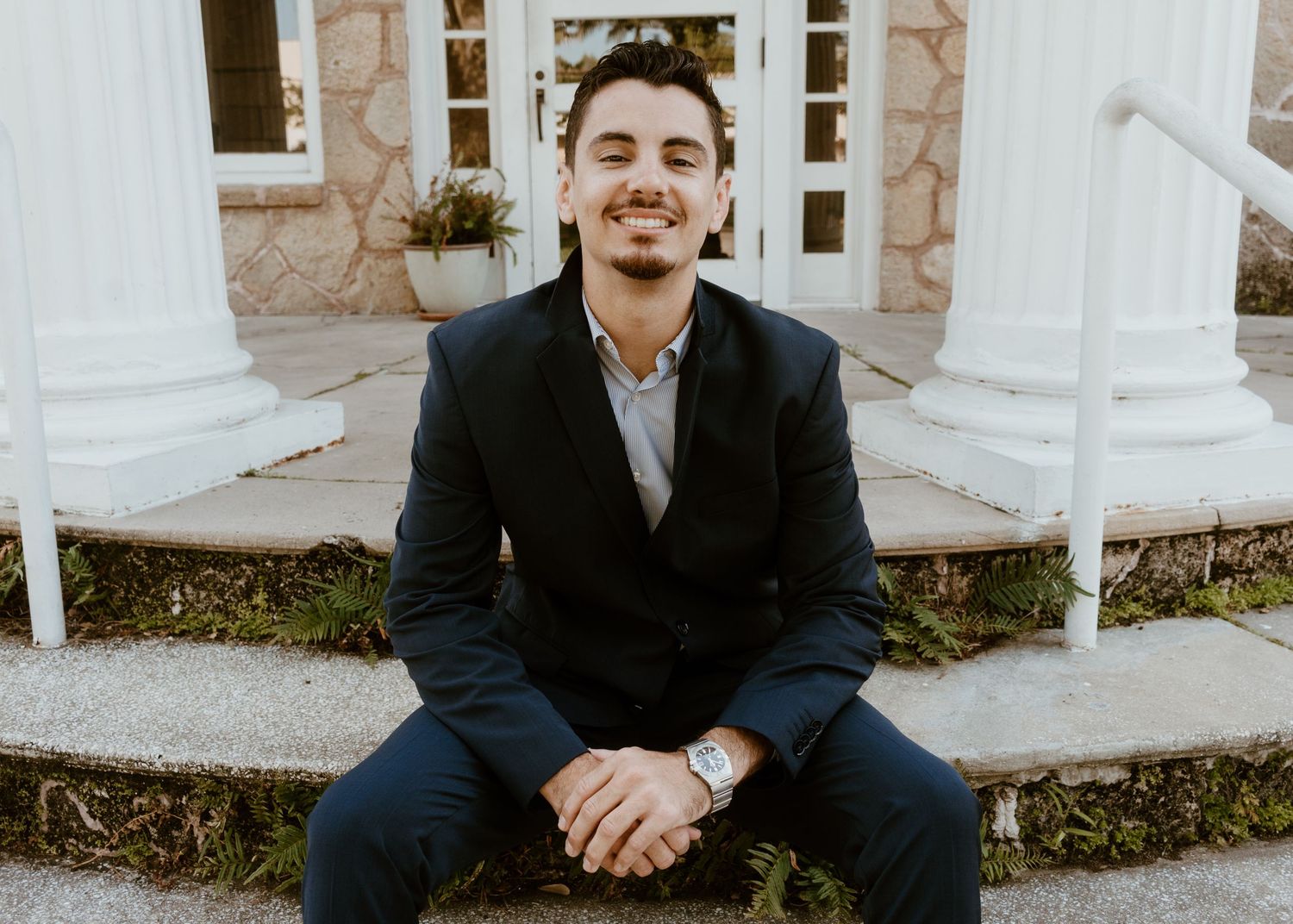 Young man in dark blazer smiling while sitting on steps in front of a building with white columns.