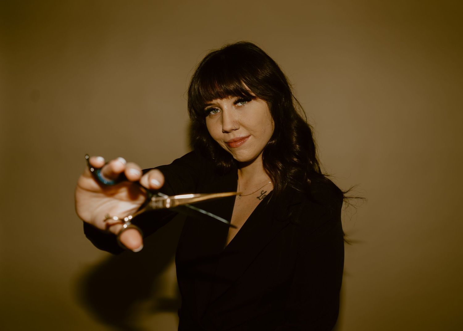 Woman with dark hair in black outfit holding scissors toward camera against neutral background.