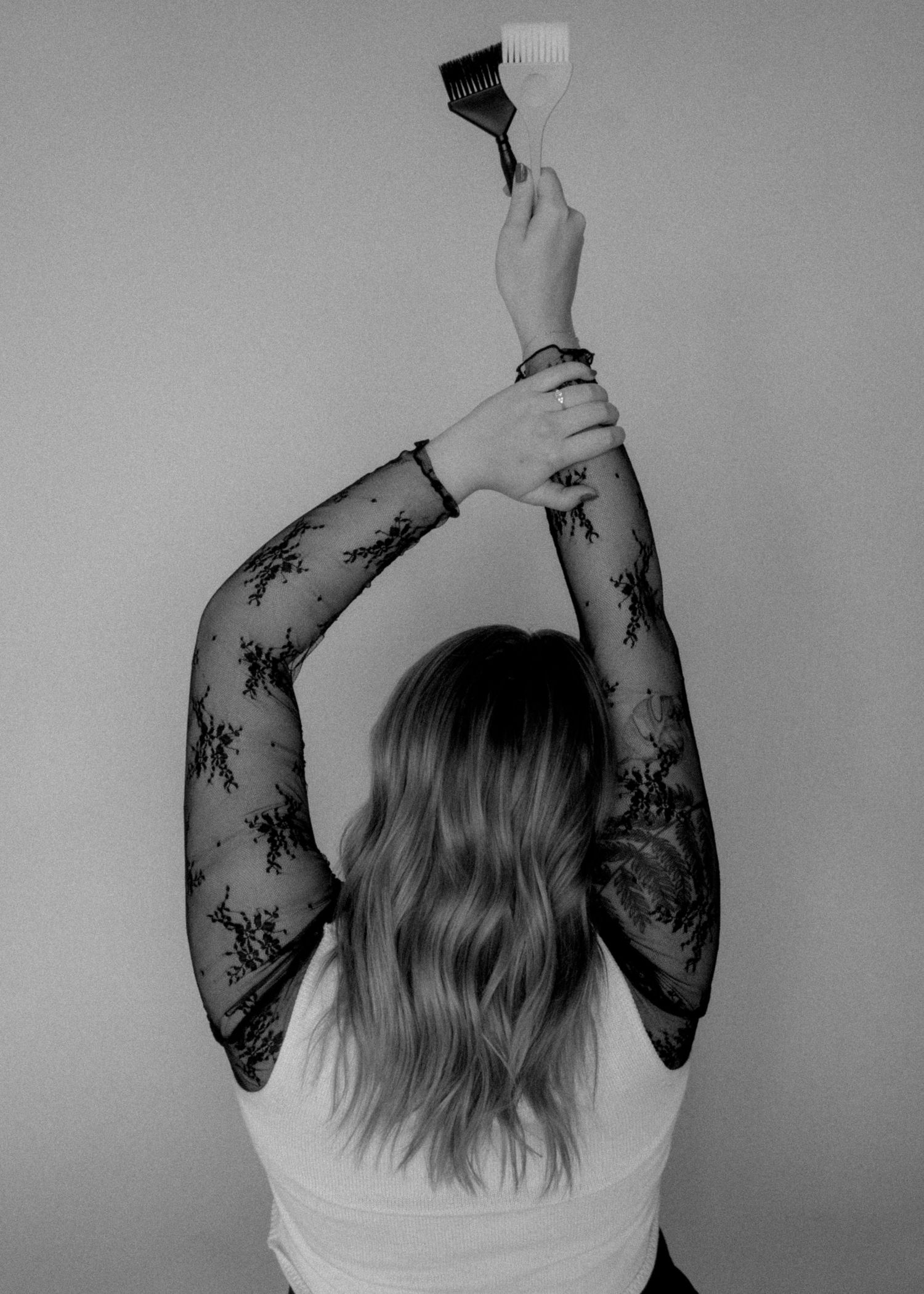 Black and white photo of a woman with long hair and lace sleeves holding a paintbrush overhead, viewed from behind.