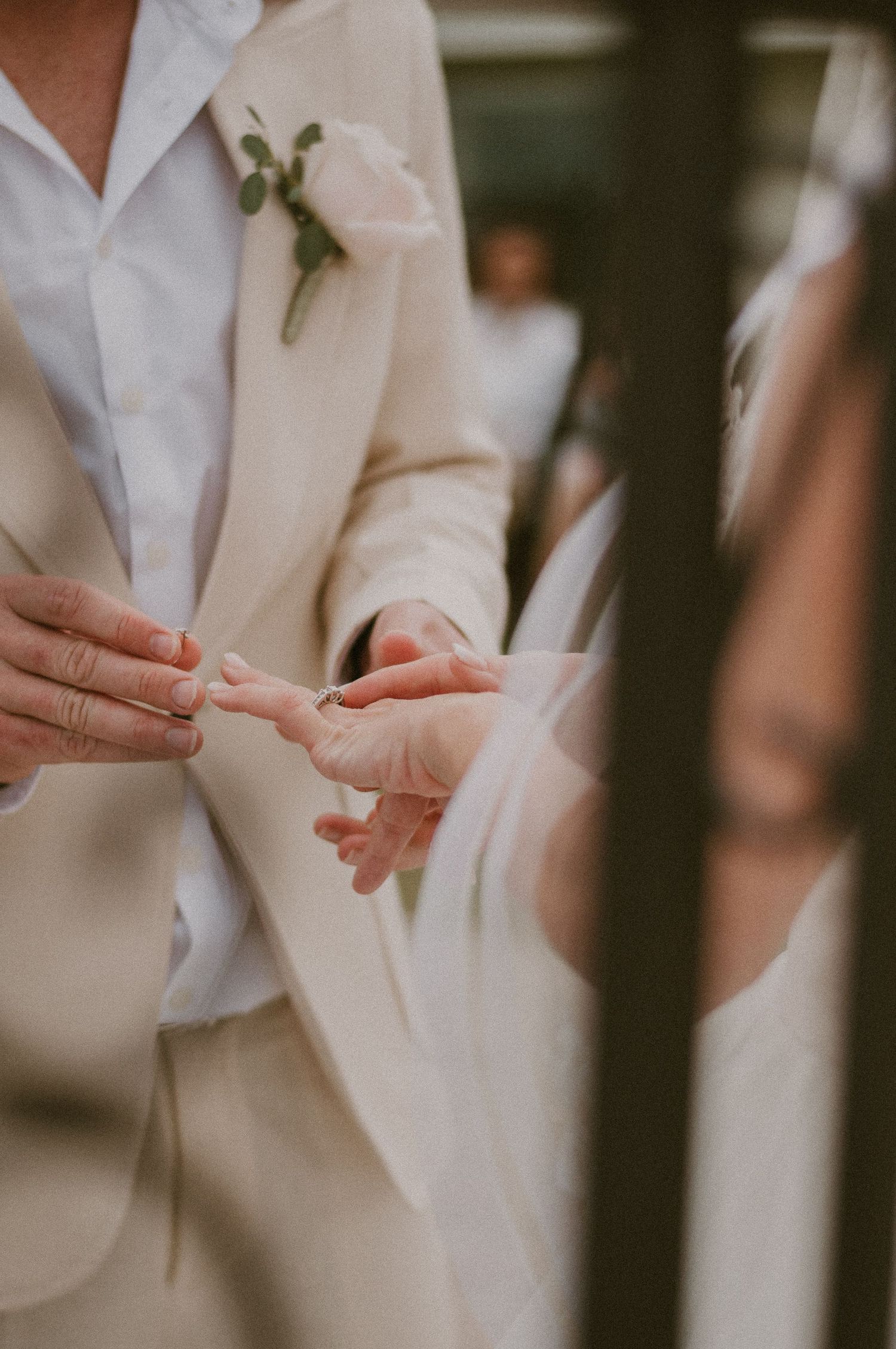 Groom in beige suit places wedding ring on brides finger during outdoor ceremony.