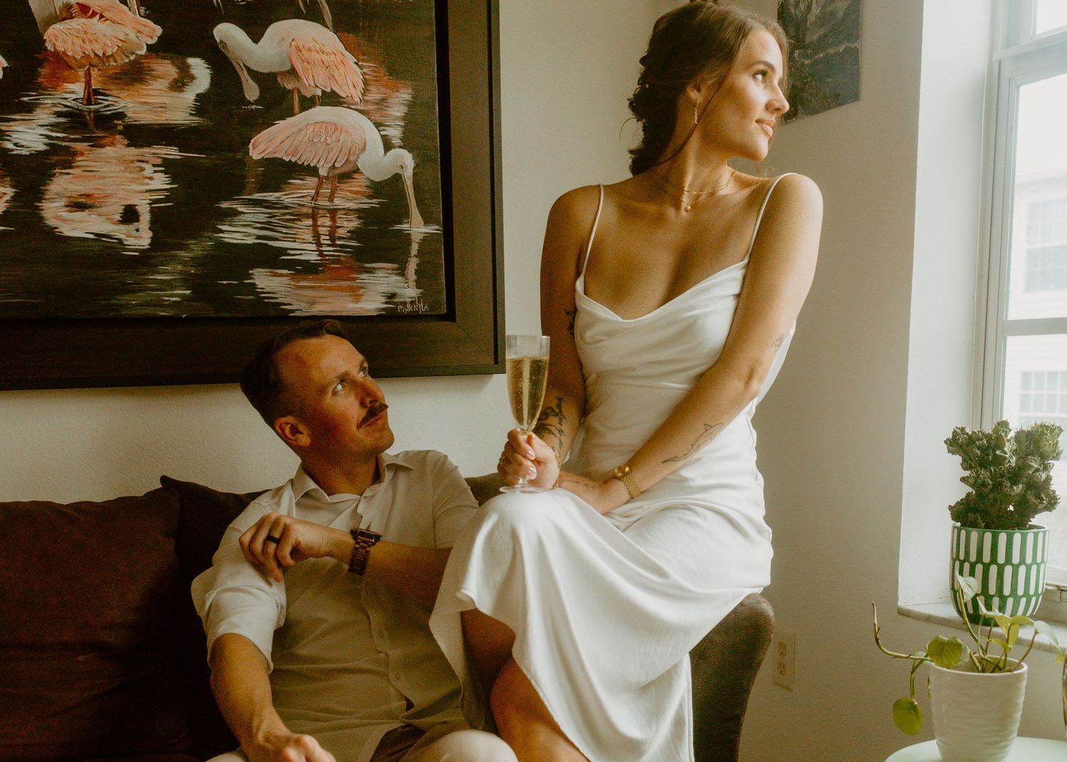 Bride in white slip dress holding champagne stands beside groom in white shirt seated on sofa near flamingo artwork.