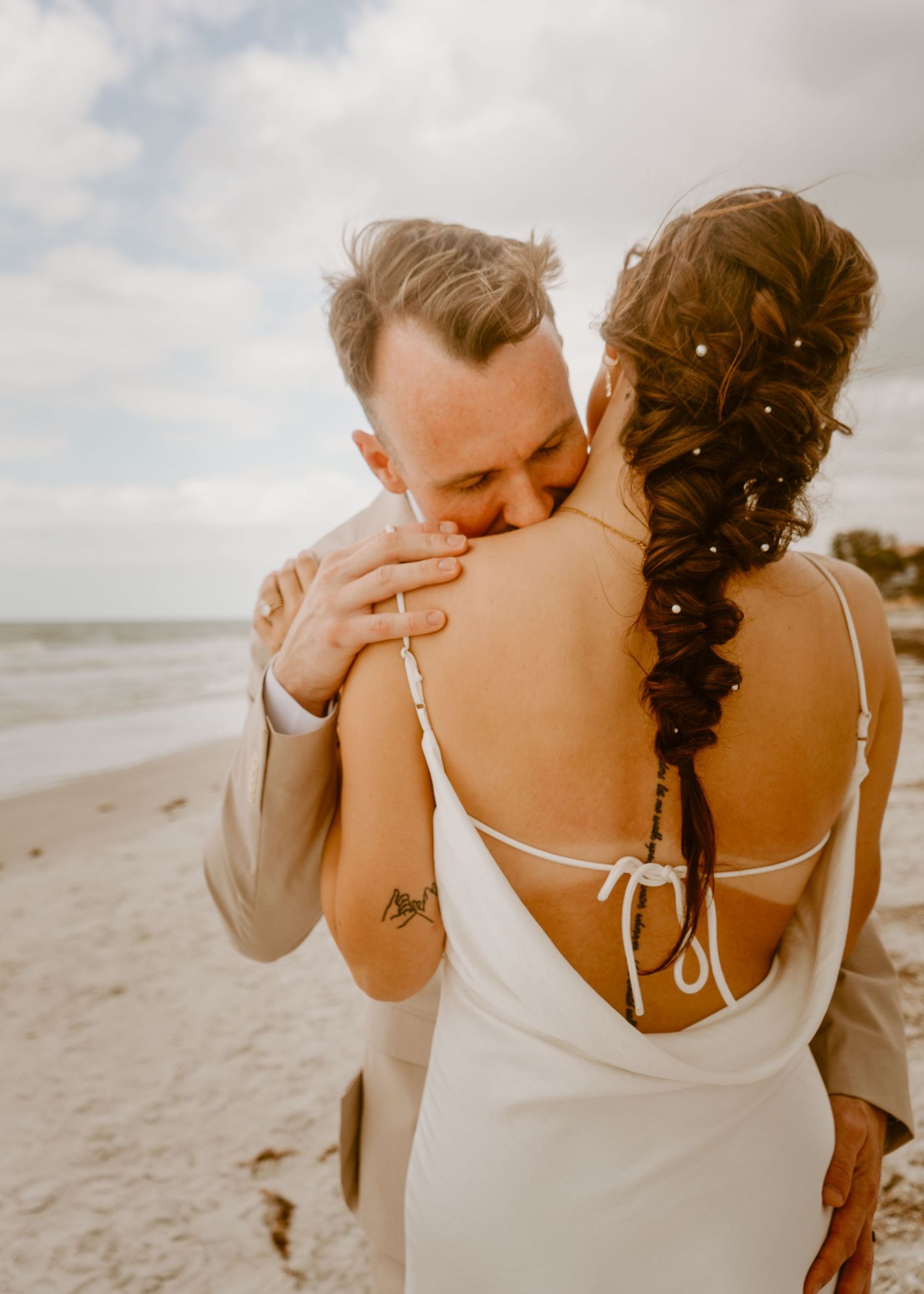 Couple embracing on beach during wedding, bride in backless white gown with braided hair, groom in tan suit.