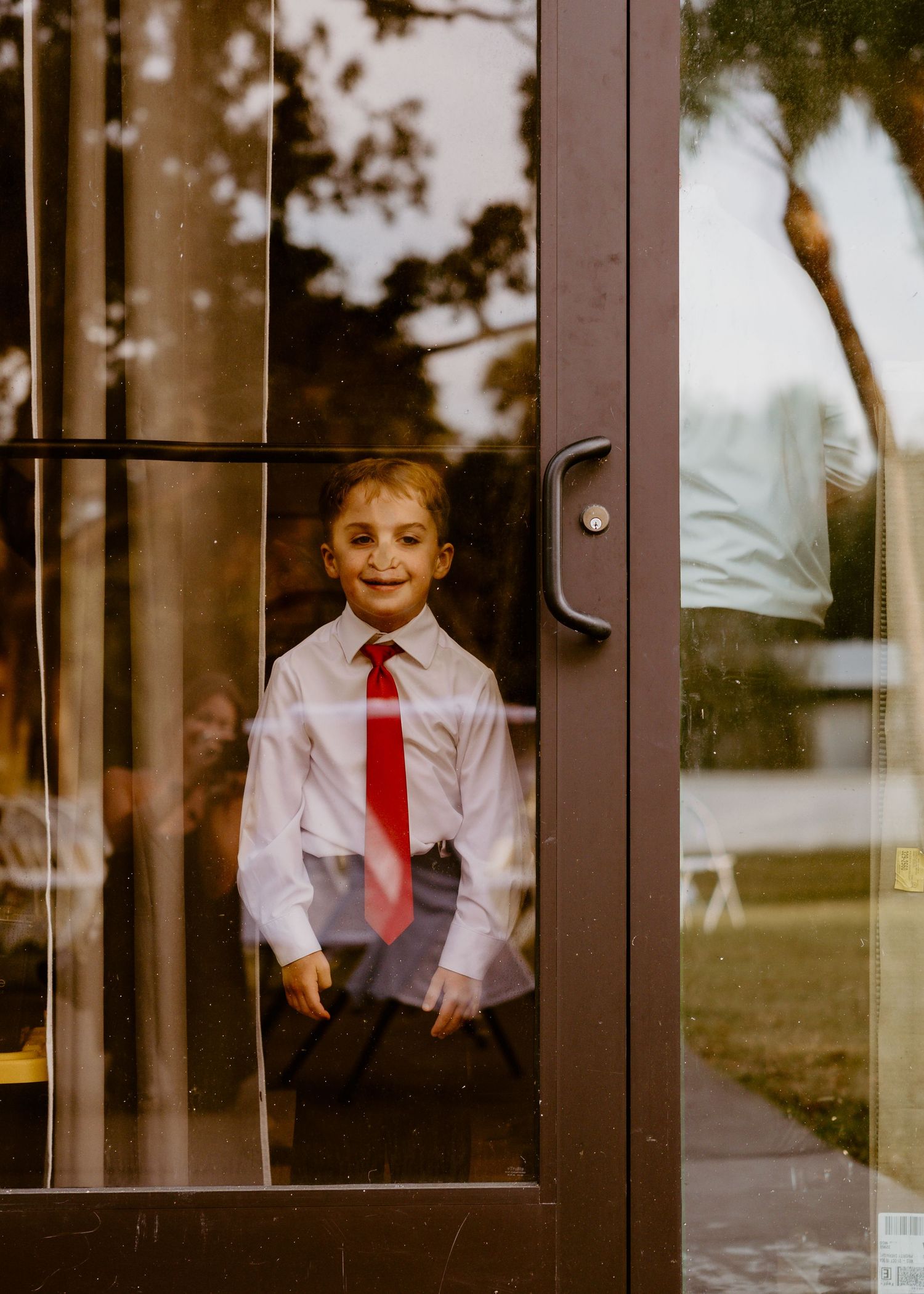 A smiling young boy in a white dress shirt and red tie peers through a glass door at an outdoor event.