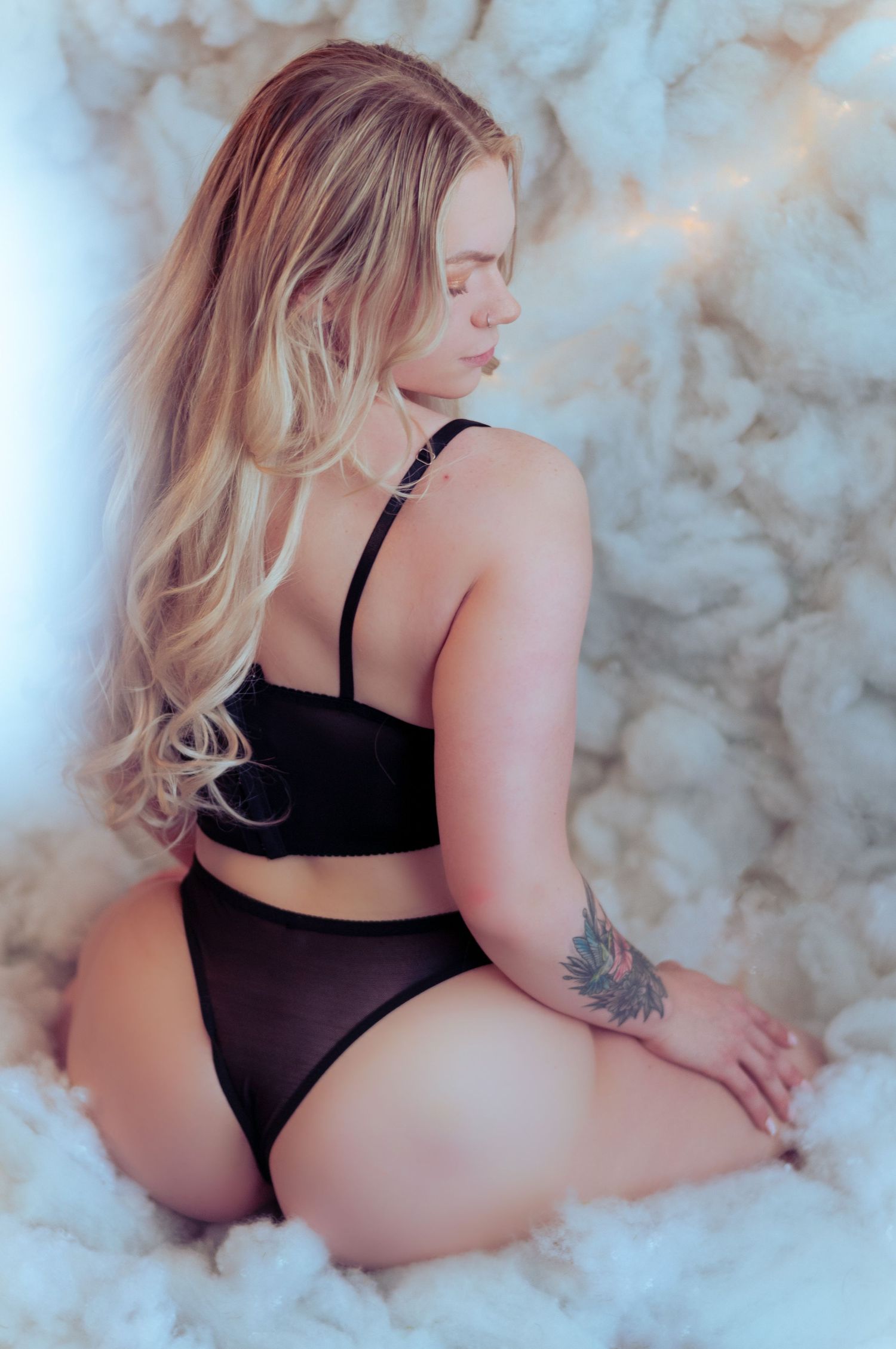 Blonde woman in black lingerie set kneeling on fluffy white surface, showing tattoo on arm.