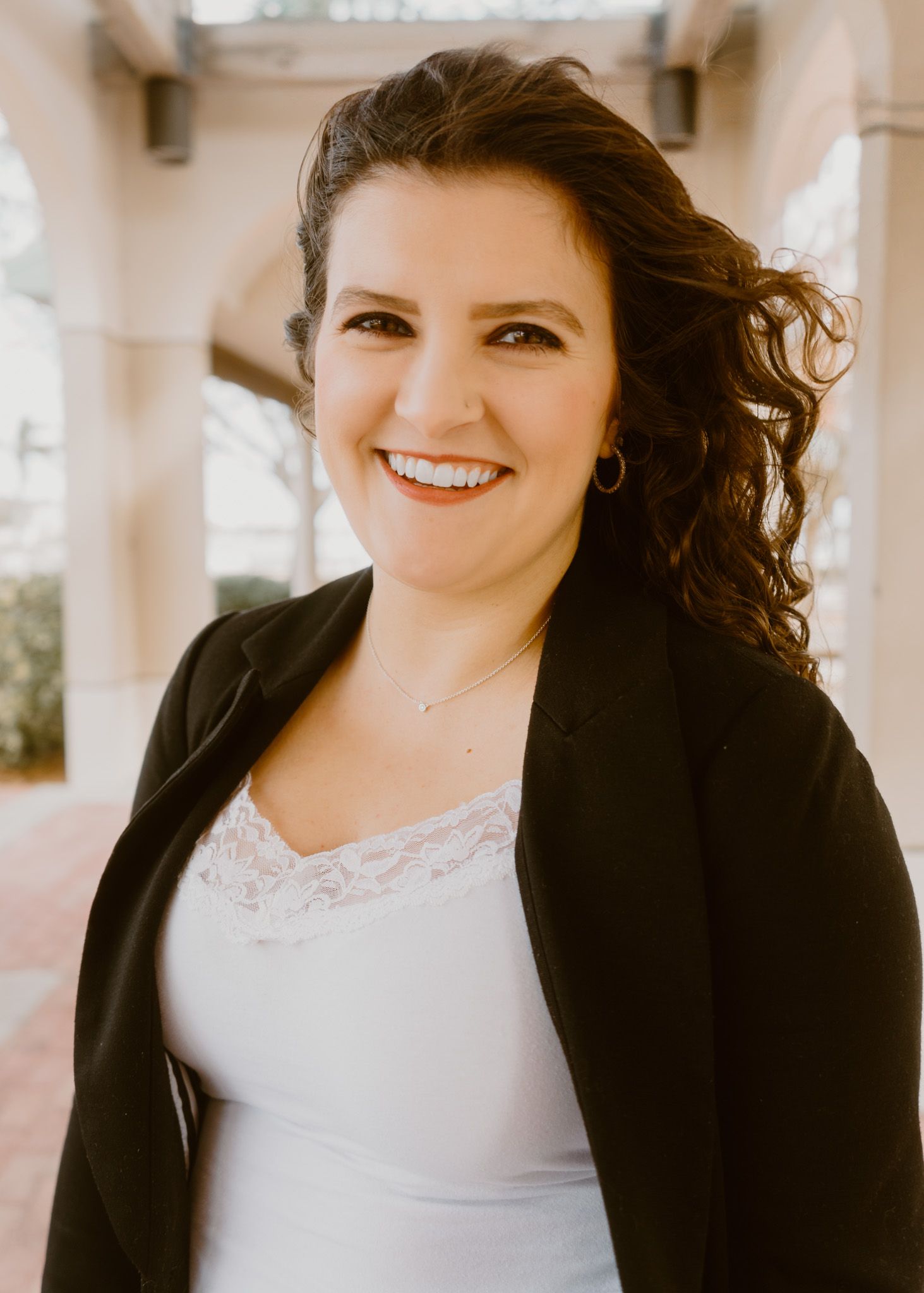Smiling woman with curly dark hair wearing a black blazer and white lace top, posing outdoors near elegant columns.