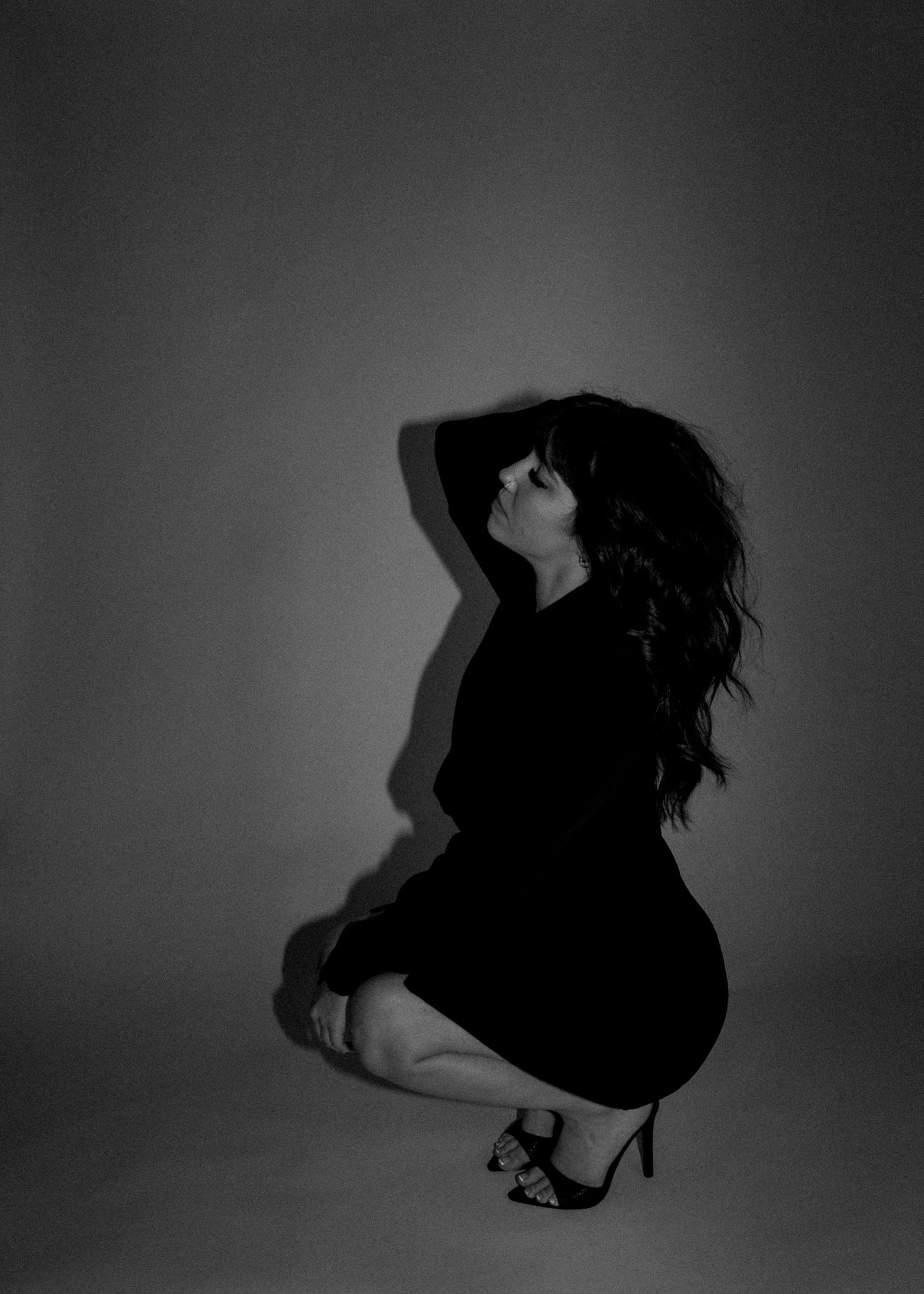 Black and white photo of a woman in a black dress and heels crouching dramatically against a studio backdrop.