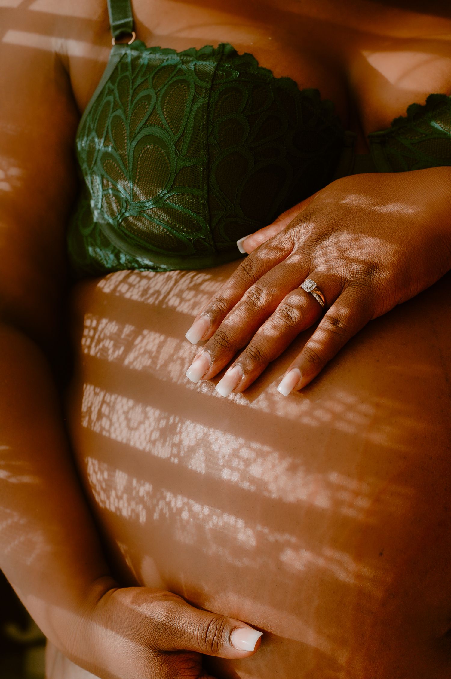 Pregnant woman in green lace bra cradling baby bump, hands adorned with diamond ring, warm light casting shadows.