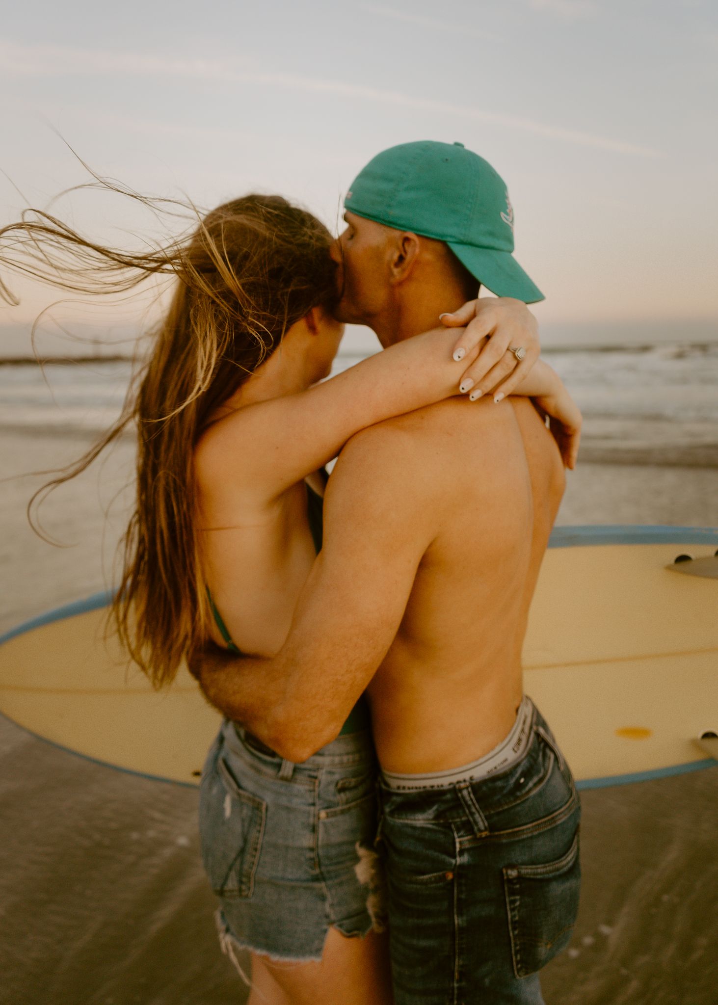 Couple kissing on beach at sunset with surfboard, woman in denim shorts with hair blowing, man in green cap.