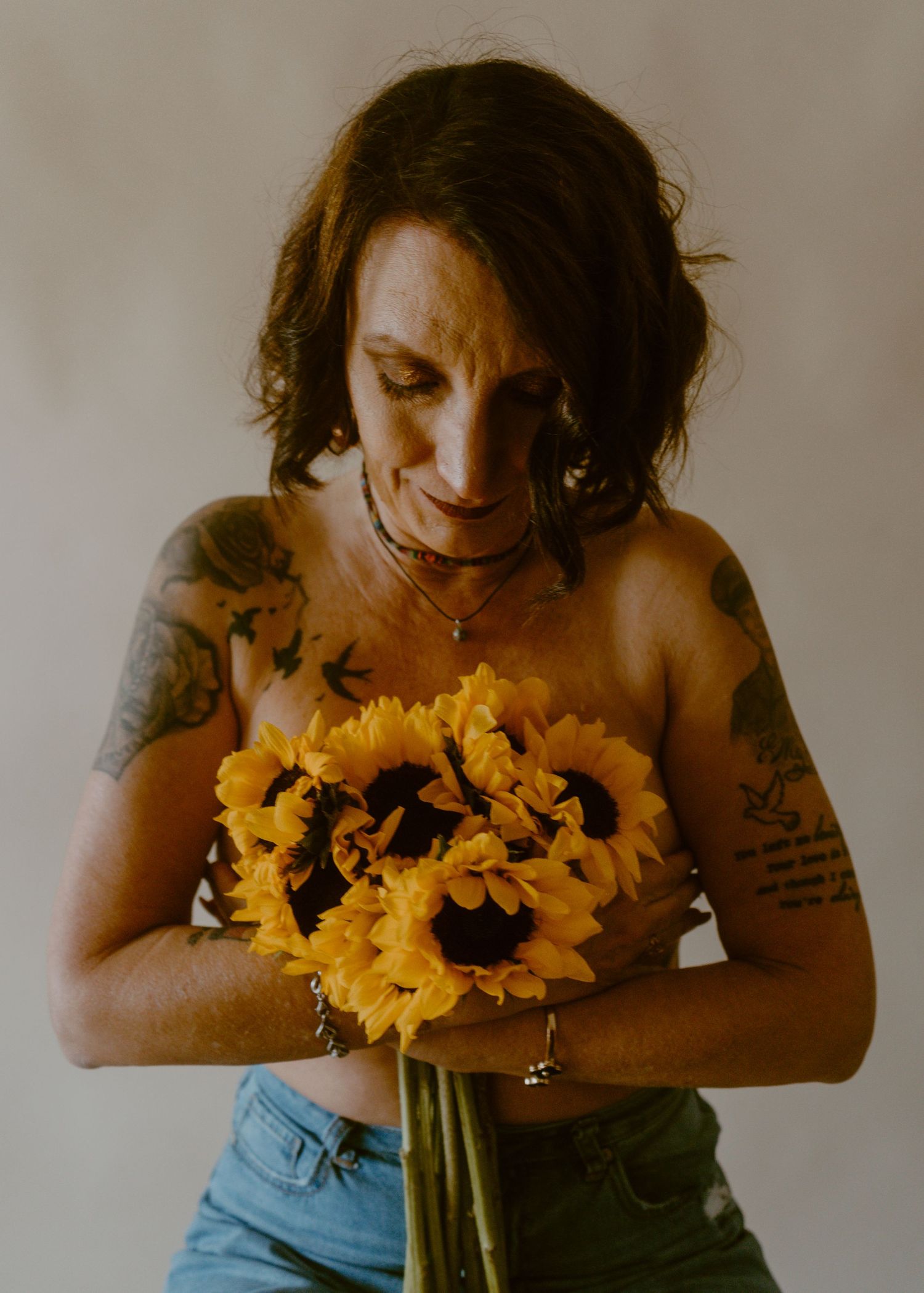 Tattooed woman holding vibrant yellow sunflowers, looking down, wearing jeans against neutral background.
