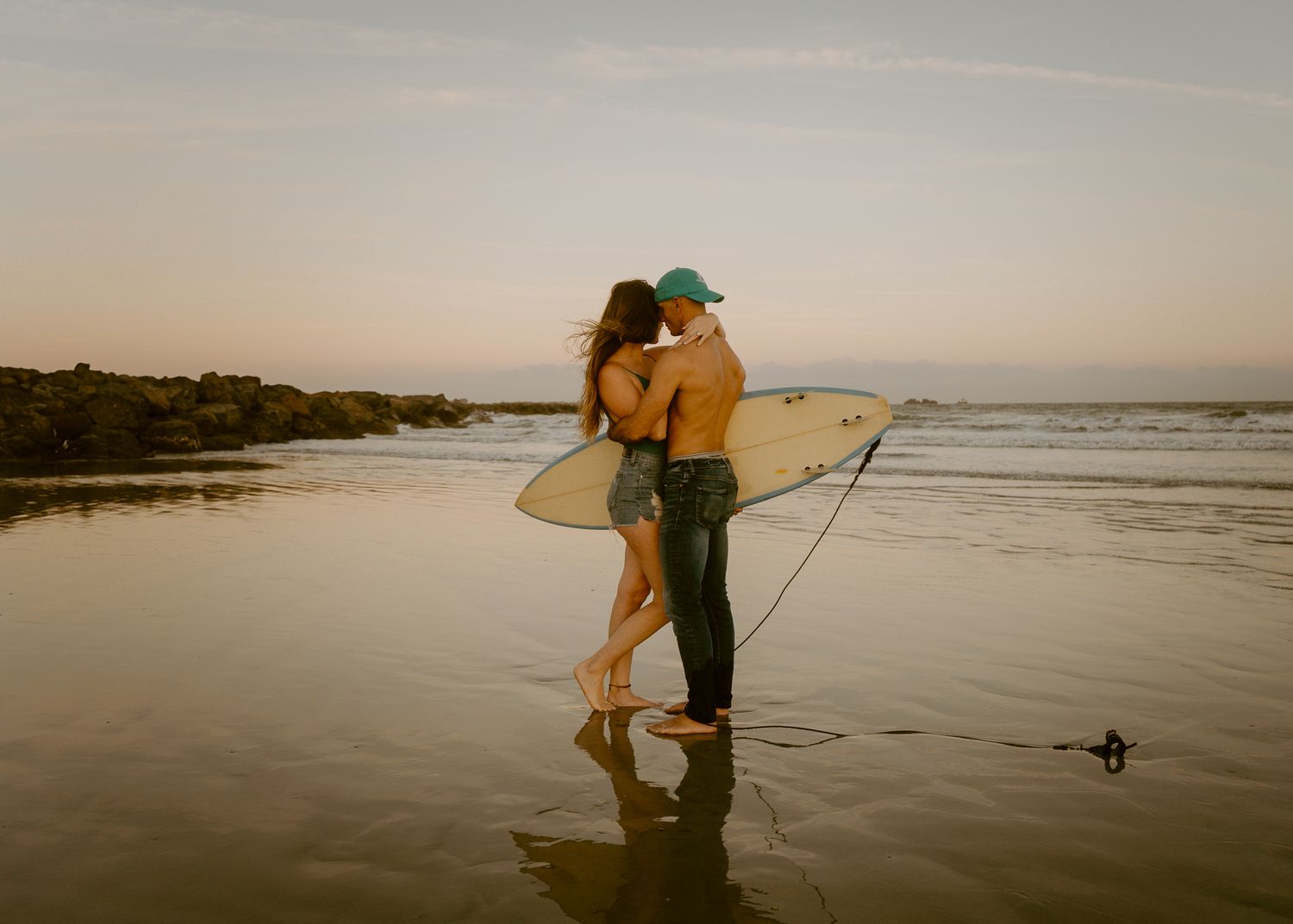 Couple kissing on beach at sunset while holding a white surfboard in shallow water.