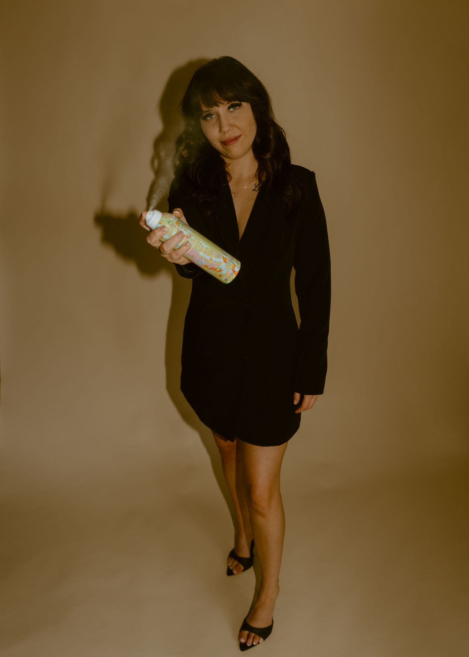 Woman in black blazer dress holds colorful hair product spray can, posing against warm brown background.