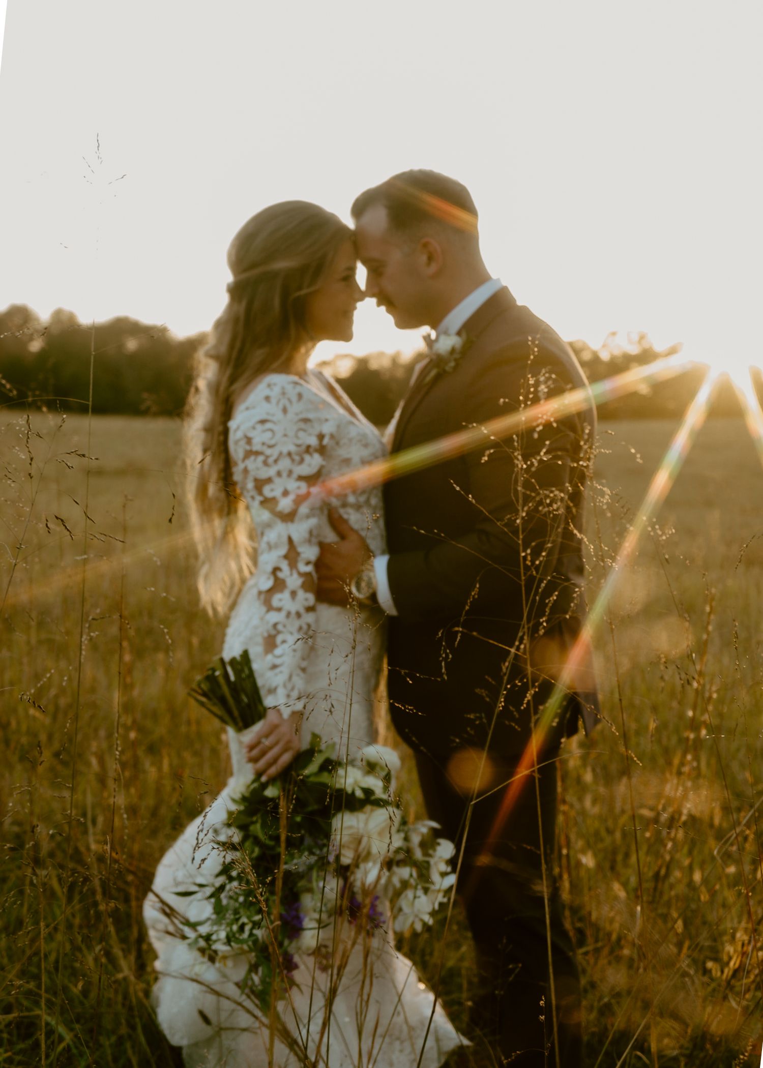 A couple shares an intimate moment at golden hour in a field, bride in lace gown holding bouquet, groom in dark suit.