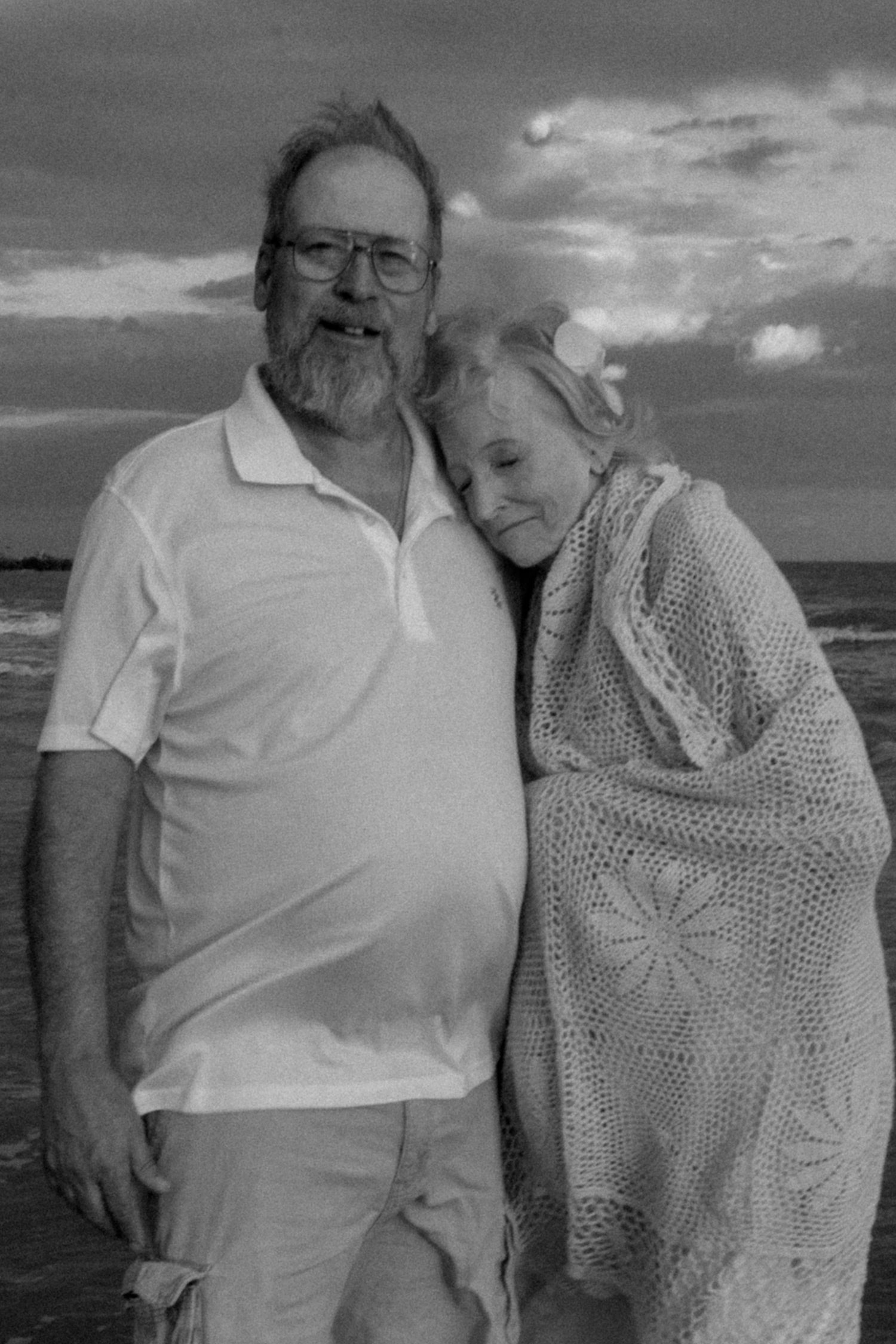 Black and white photo of an older couple smiling together on a beach, woman resting head on mans shoulder.