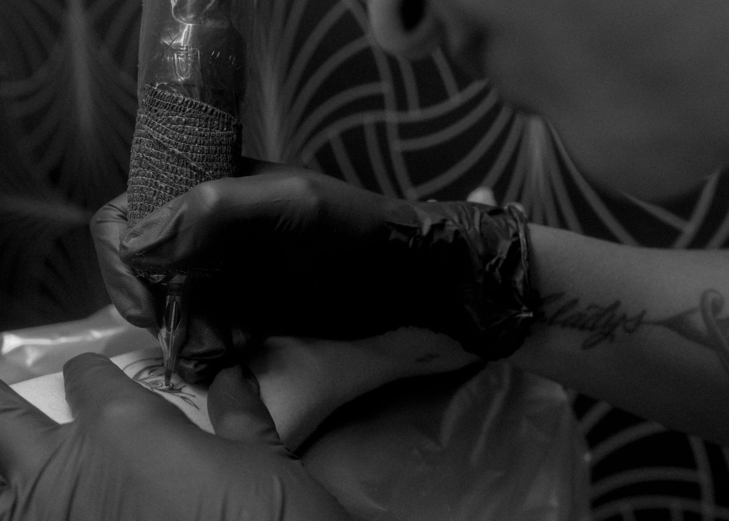Tattoo artist in black gloves carefully applying ink to a client's hand in a black and white photo.
