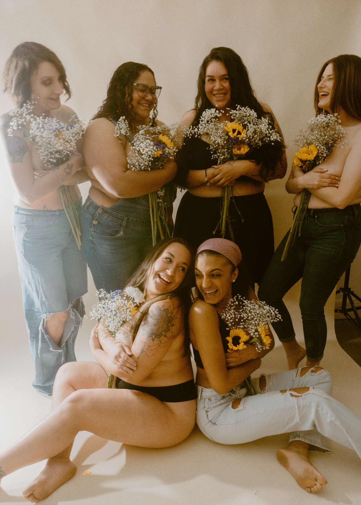 Six diverse women posing together holding sunflower and baby's breath bouquets in a bright studio setting.