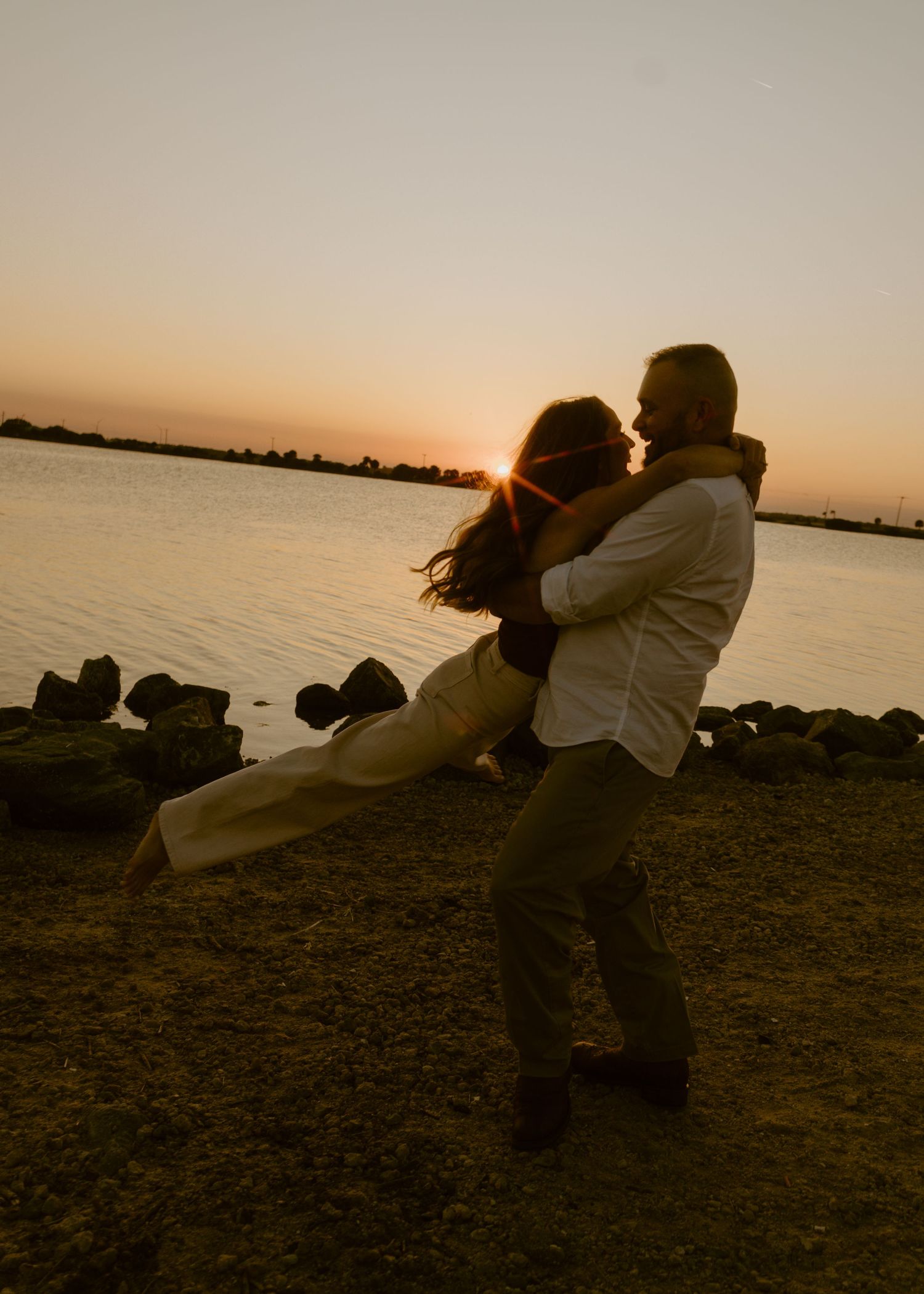 Couple embracing at sunset by rocky waterfront, silhouetted against golden sky.