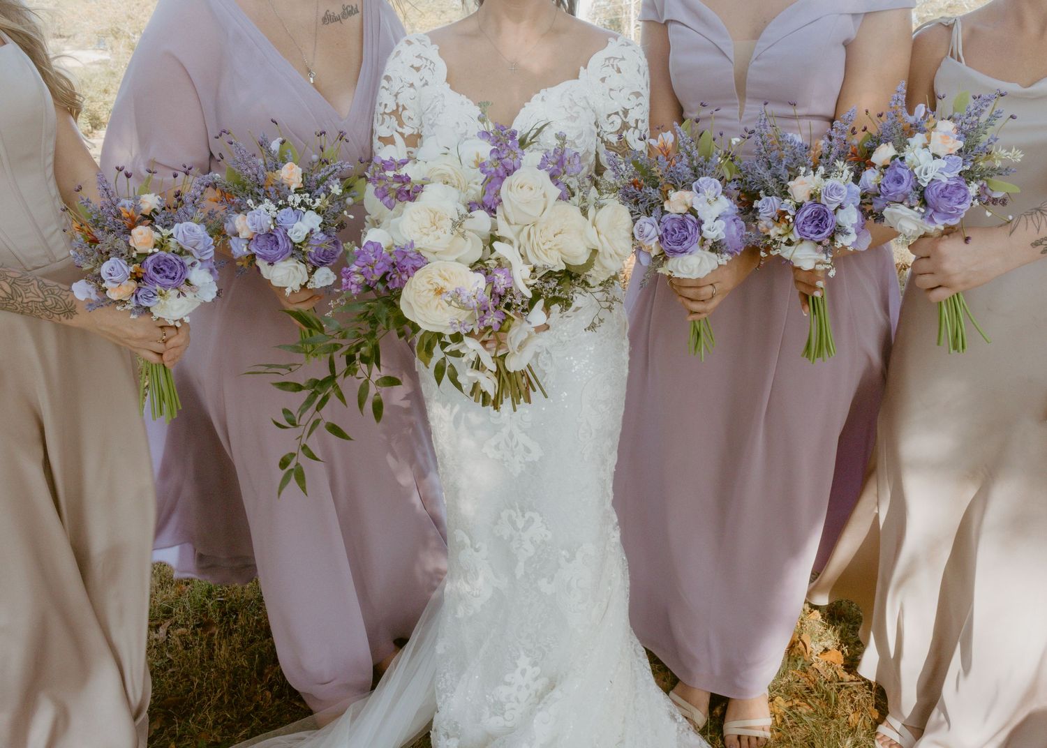 Bride in lace gown with bridesmaids in lavender dresses holding purple and white bouquets.