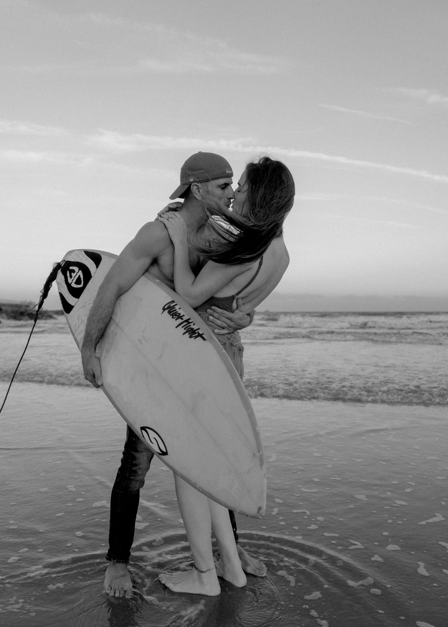 Black and white photo of a couple kissing on the beach while holding a surfboard in the ocean waves.