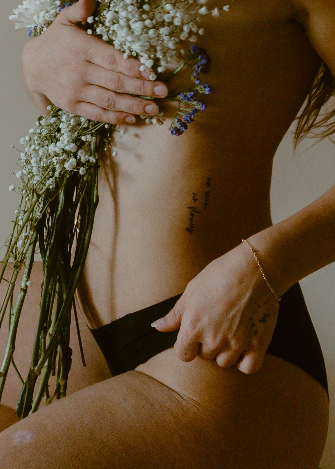 Woman in black bikini holding wildflowers with a script tattoo on her ribcage and a delicate gold bracelet.