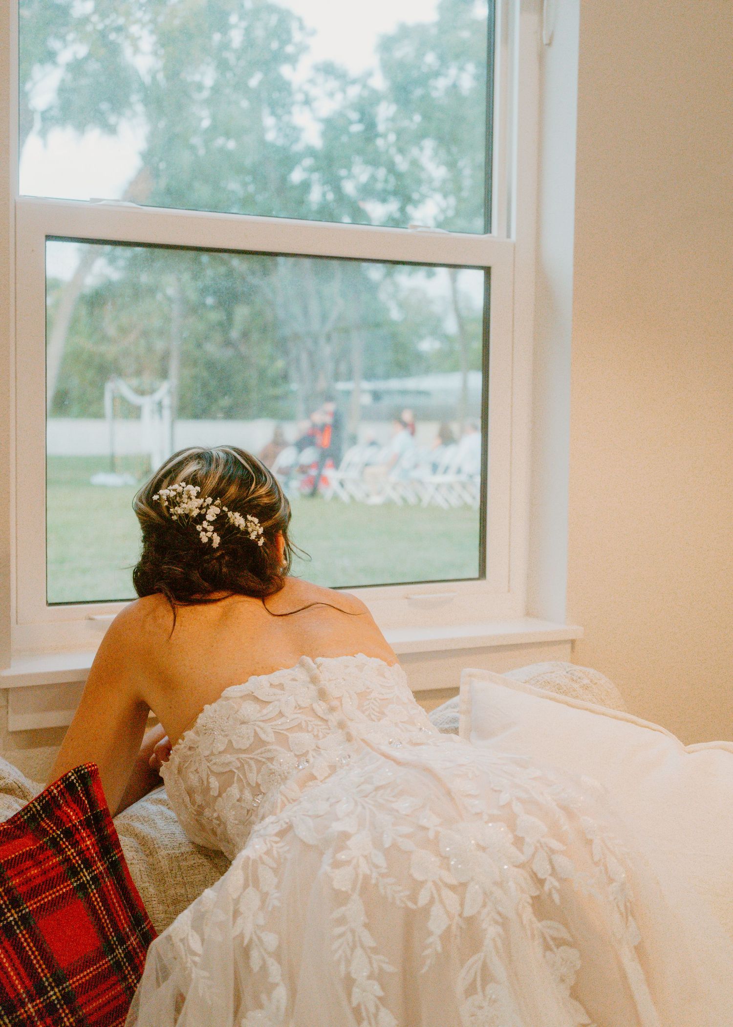 Bride in lace wedding gown with floral hair piece gazing out window at outdoor ceremony setup.