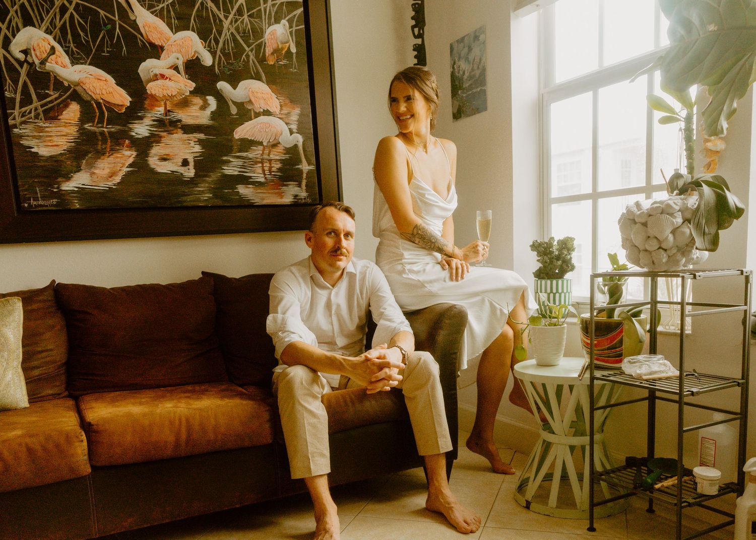 Couple relaxing in stylish living room with flamingo wall art, white outfits, champagne, and tropical plants by window.
