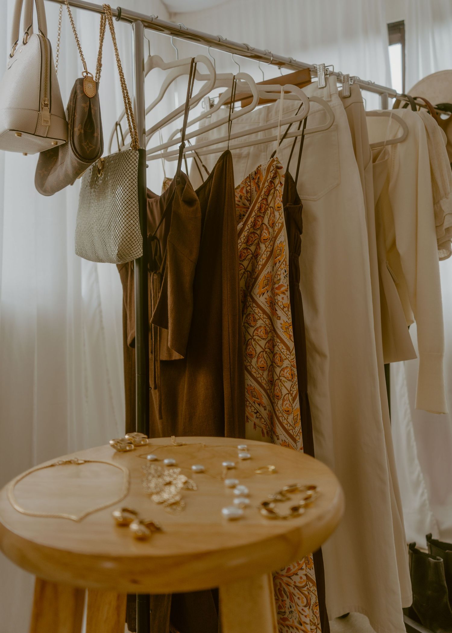 A clothing rack with neutral-toned garments and handbags, beside a wooden stool displaying gold jewelry and pearls.