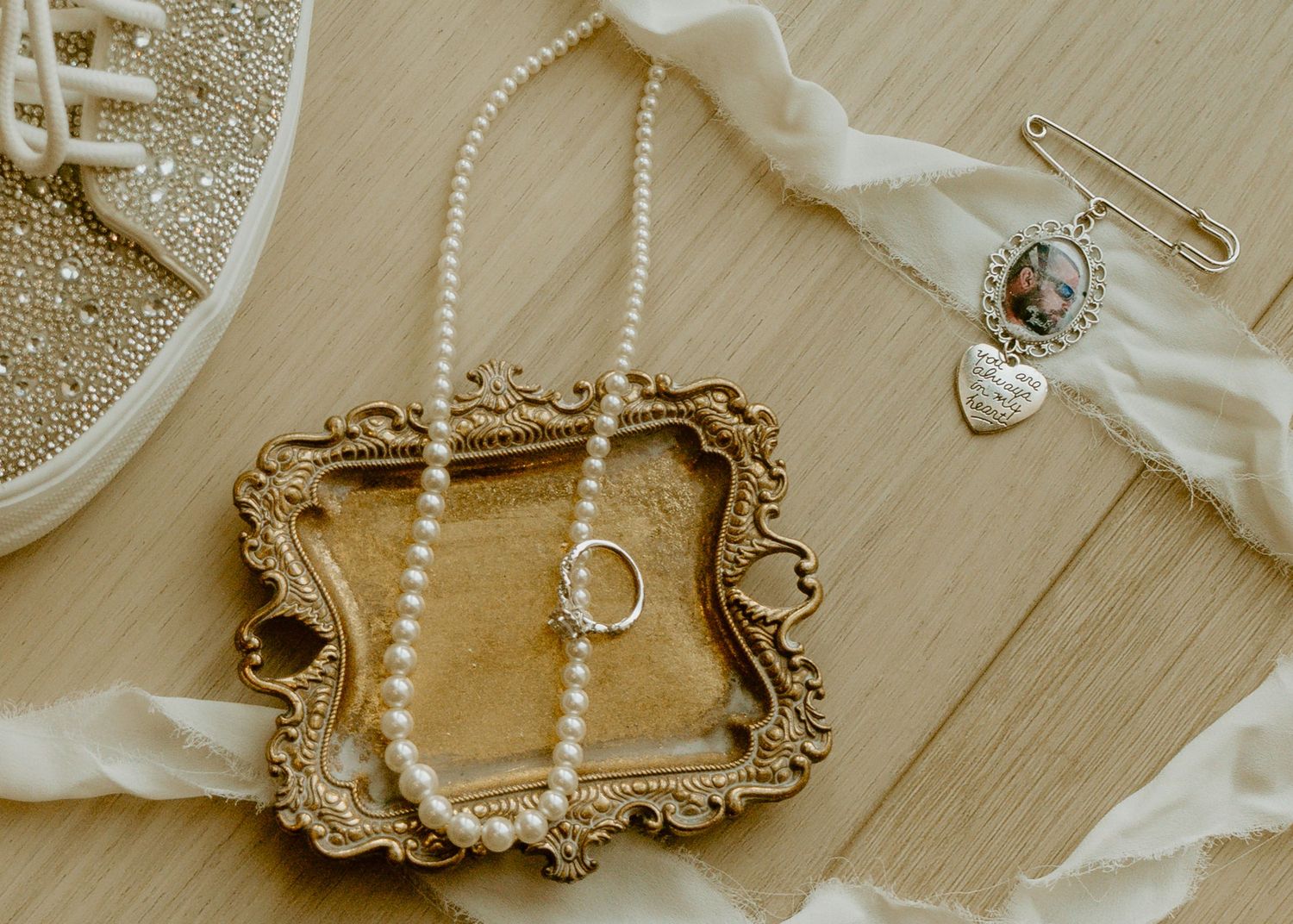 Elegant bridal flat lay with pearl necklace, gold ring, ornate frame, and heart charm on wooden surface.