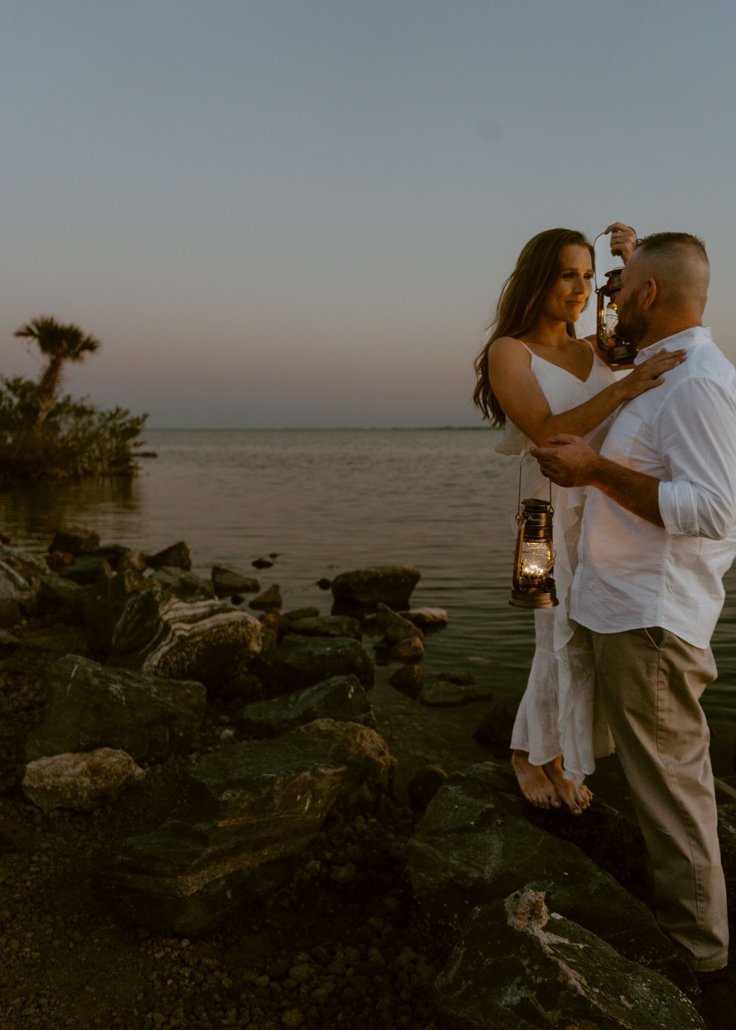 Couple embracing at sunset on rocky shoreline, woman holding vintage lantern, wearing white outfits near calm water.