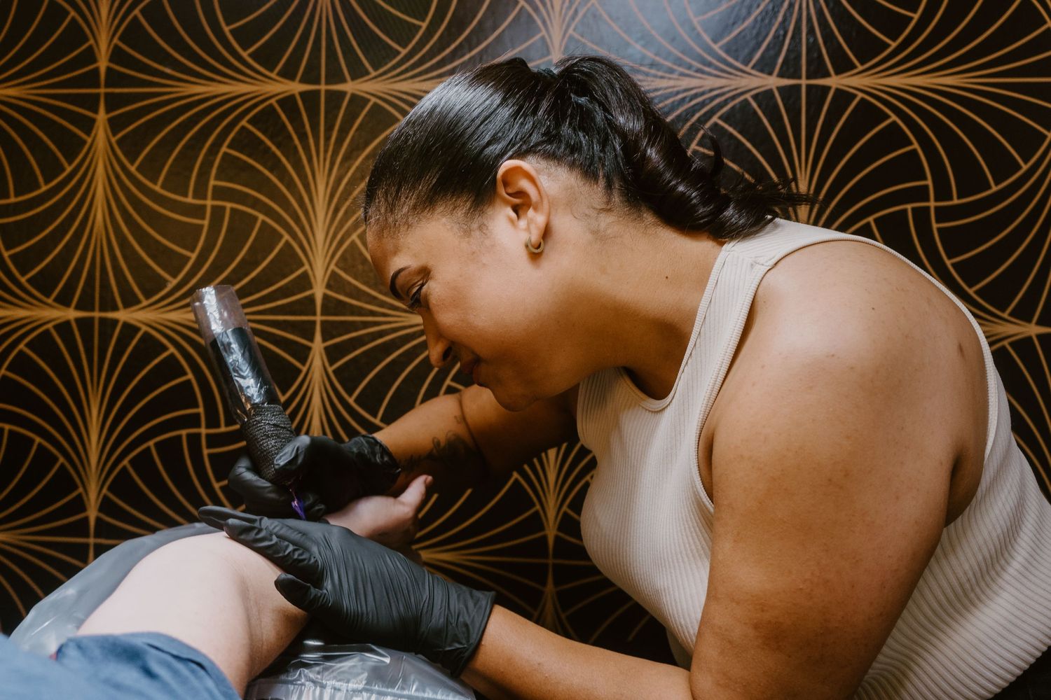 Female tattoo artist wearing black gloves carefully tattooing a client's arm in a stylish studio.
