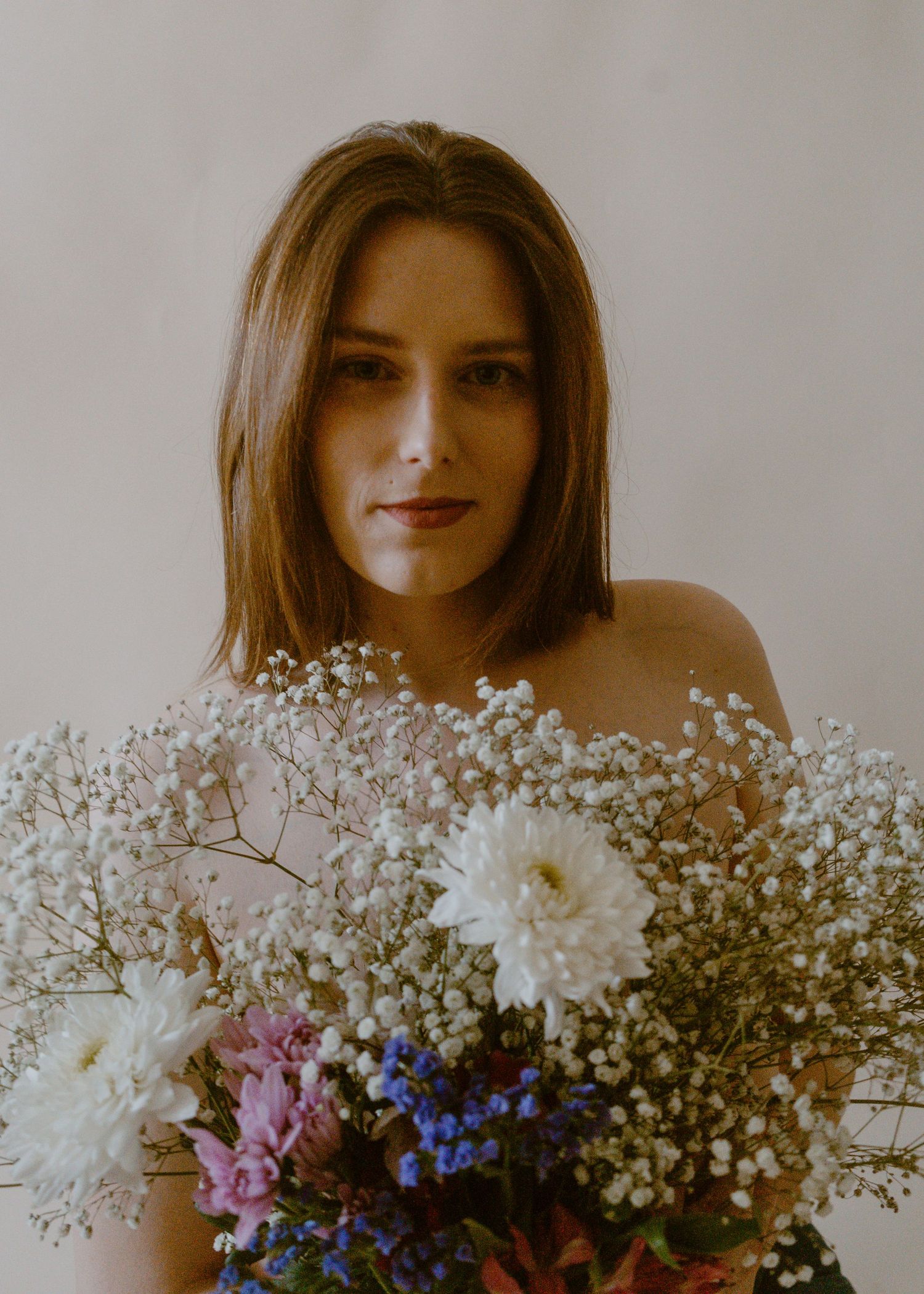 Young woman with brown hair holding a lush bouquet of white, pink, and blue wildflowers against a neutral background.