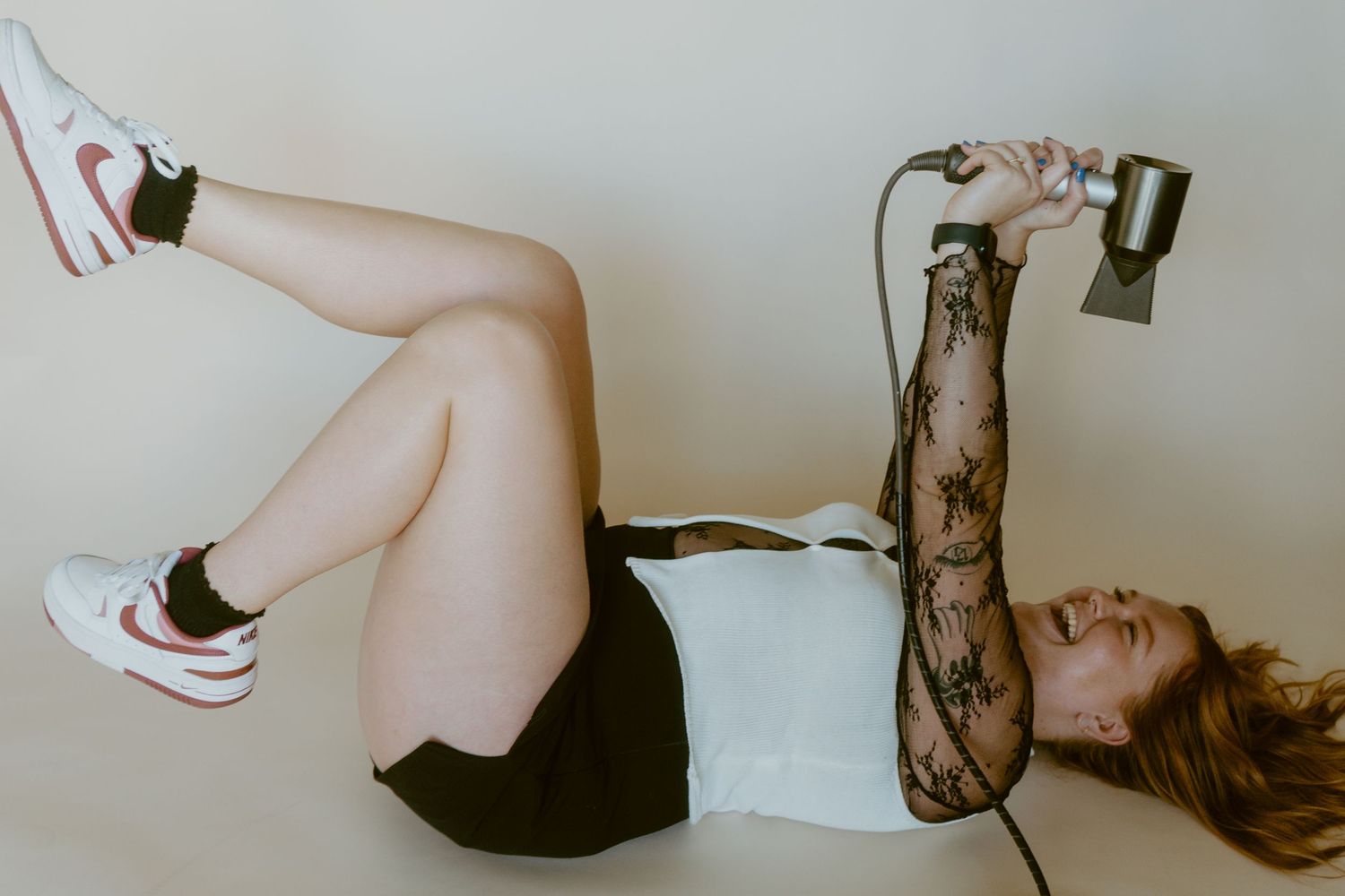 Woman lying on floor in white tank, black skirt, and Air Jordans, holding a microphone above her head.
