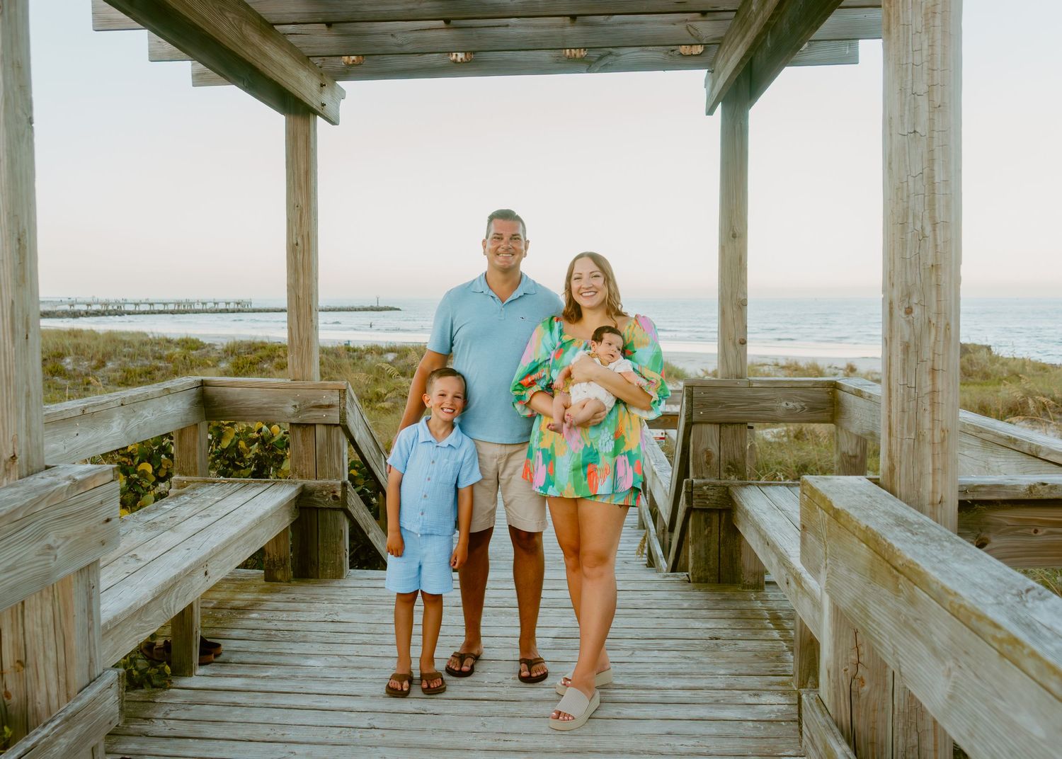 Family of four posing on a wooden beach boardwalk at sunset, parents with two young children near the ocean.