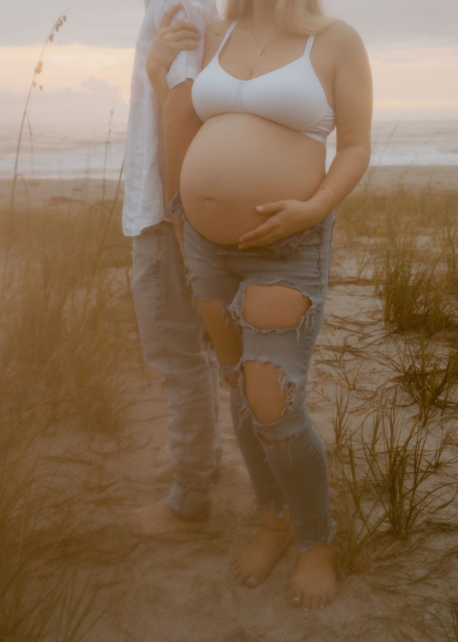 Pregnant woman in white bralette and ripped jeans cradling baby bump on sandy beach at sunset.