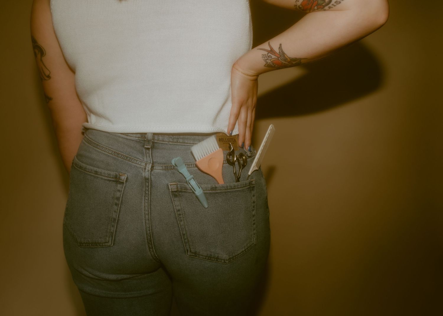Tattooed woman in white tank top and gray jeans with makeup brushes and tools in back pocket.