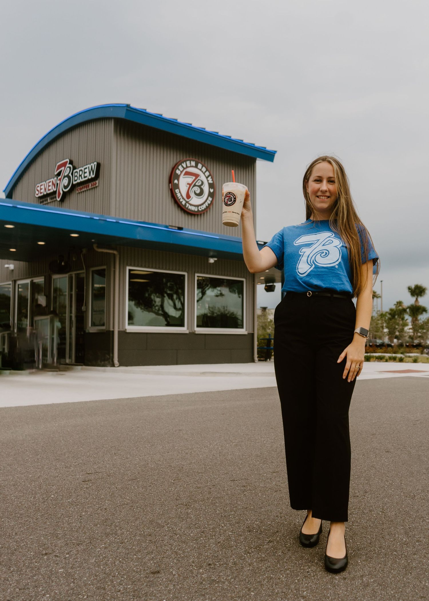 Woman holding iced coffee drink outside Seven Brew Coffee shop, wearing blue graphic tee and black maxi skirt.