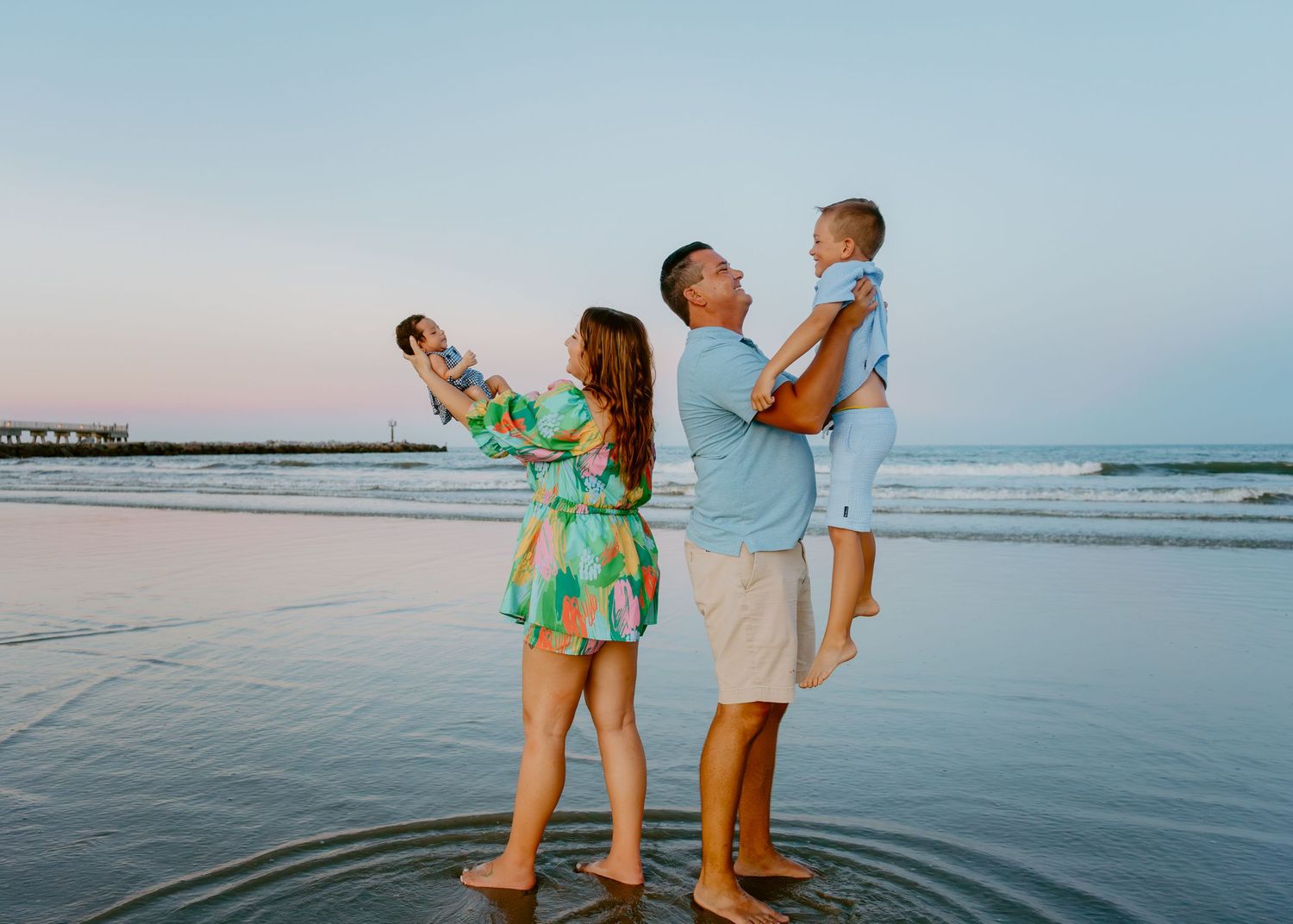 Family of three standing on a beach at sunset, parents holding toddler while woman photographs the ocean view.