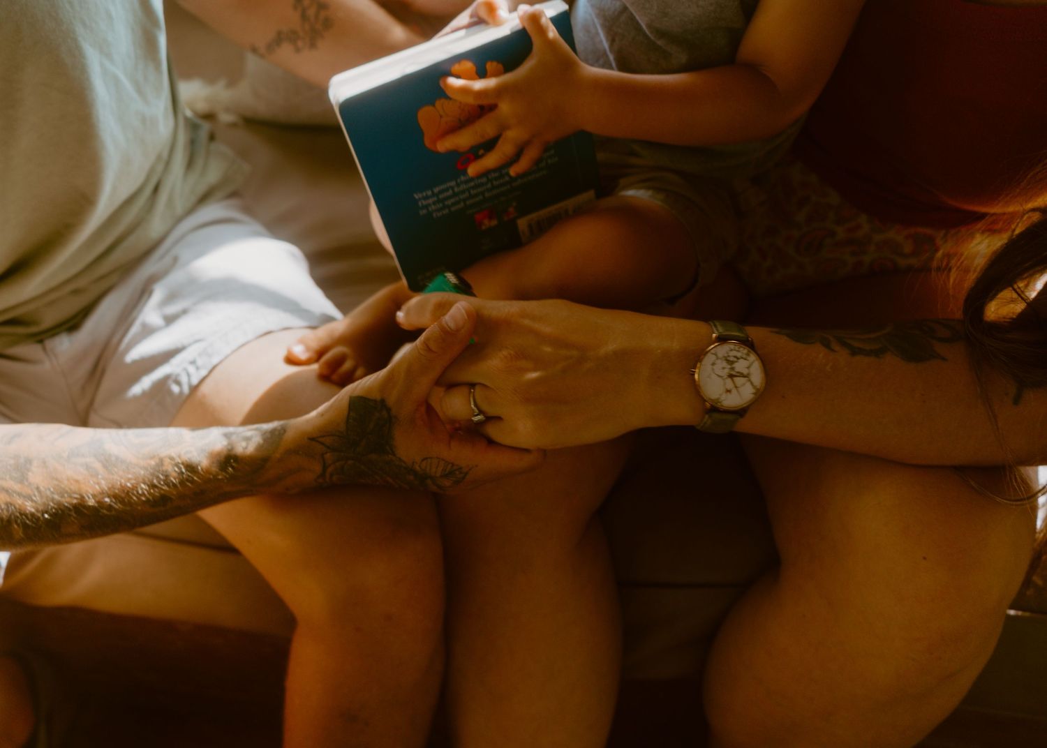 Two adults with tattoos hold hands while a child reads a teal book, sharing a cozy family reading moment.