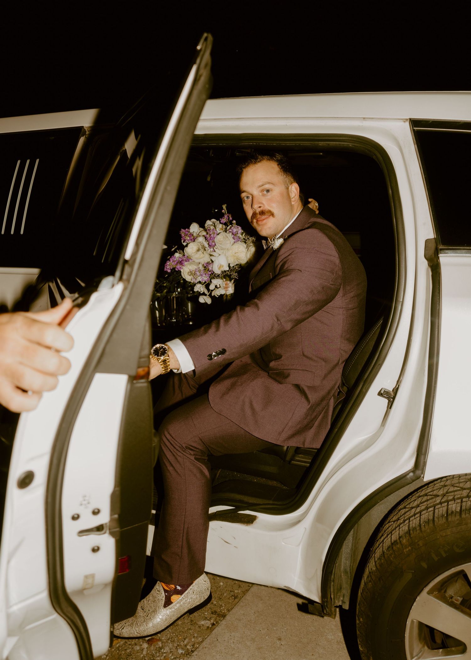 Man in brown suit holding bouquet exits white SUV at night, wearing loafers and a watch.