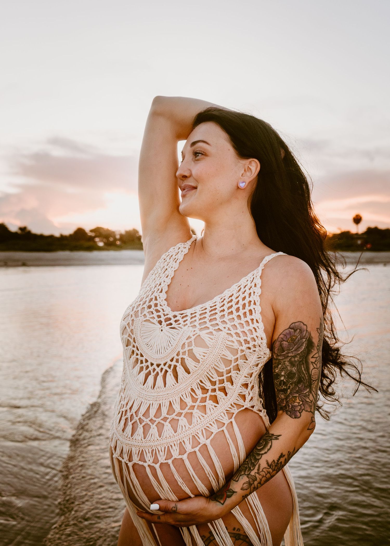 Pregnant woman in crochet dress with tattoos holding belly at sunset by the water.