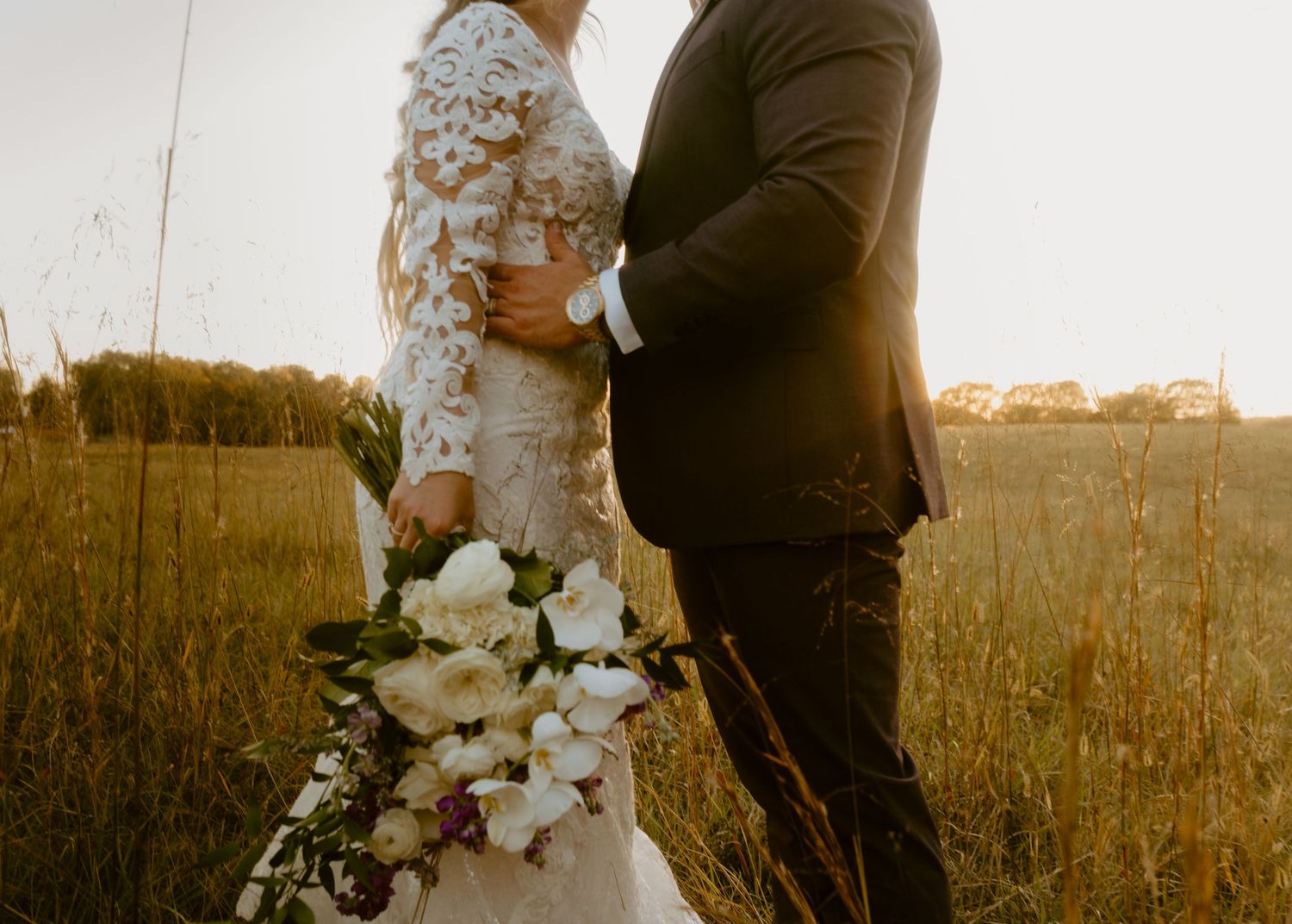 Bride and groom embracing in golden sunset field, bride holding white floral bouquet, wearing lace gown and black suit.