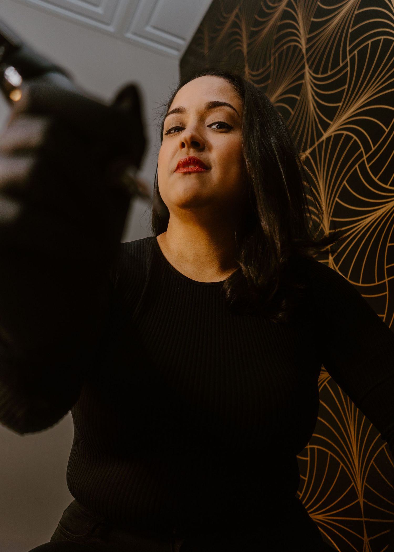 Woman in black ribbed top poses confidently against gold art deco patterned wallpaper with dramatic lighting.
