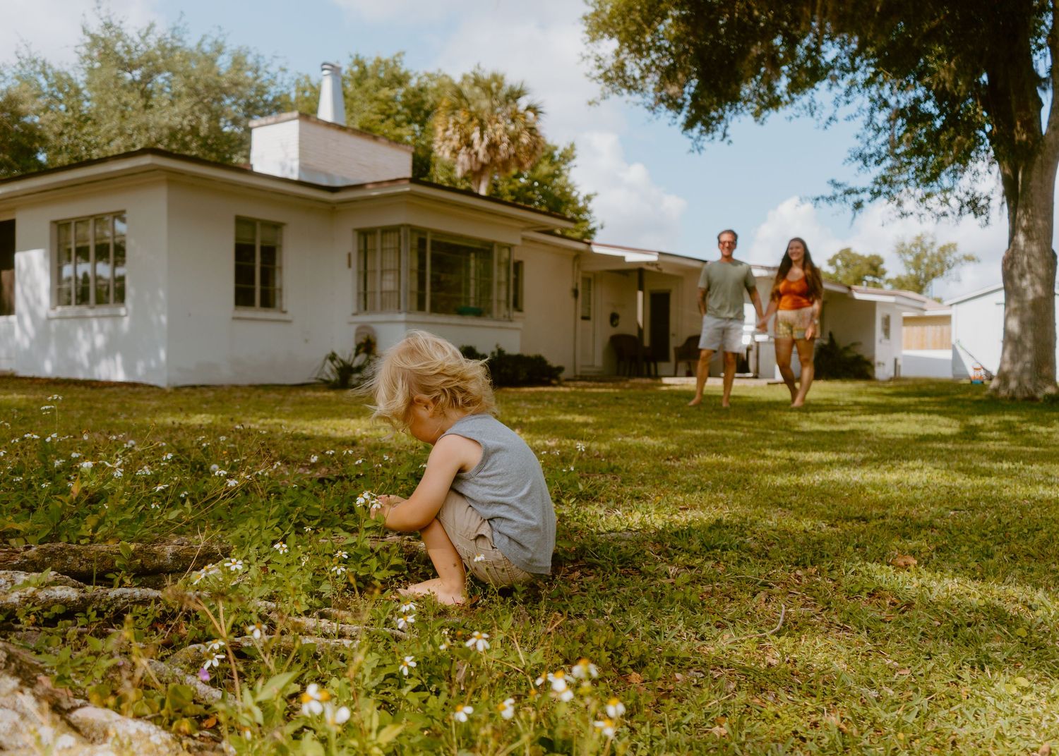 A toddler plays in green grass while parents walk toward a white ranch-style home on a sunny day.