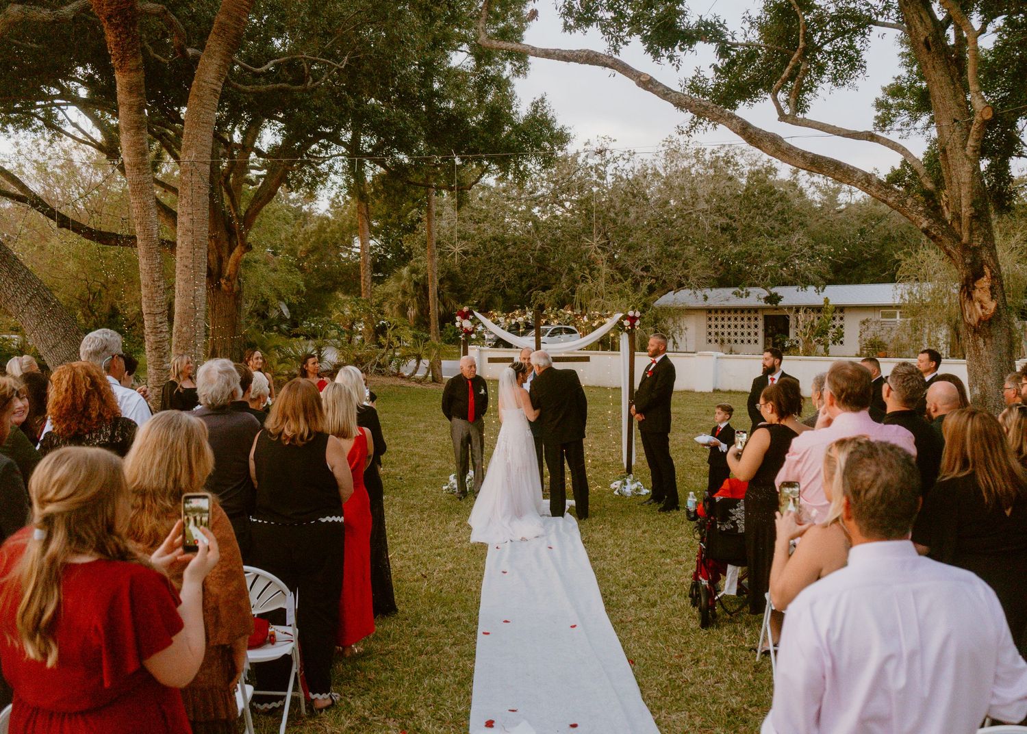 Outdoor wedding ceremony under large oak trees with guests seated along white aisle runner.