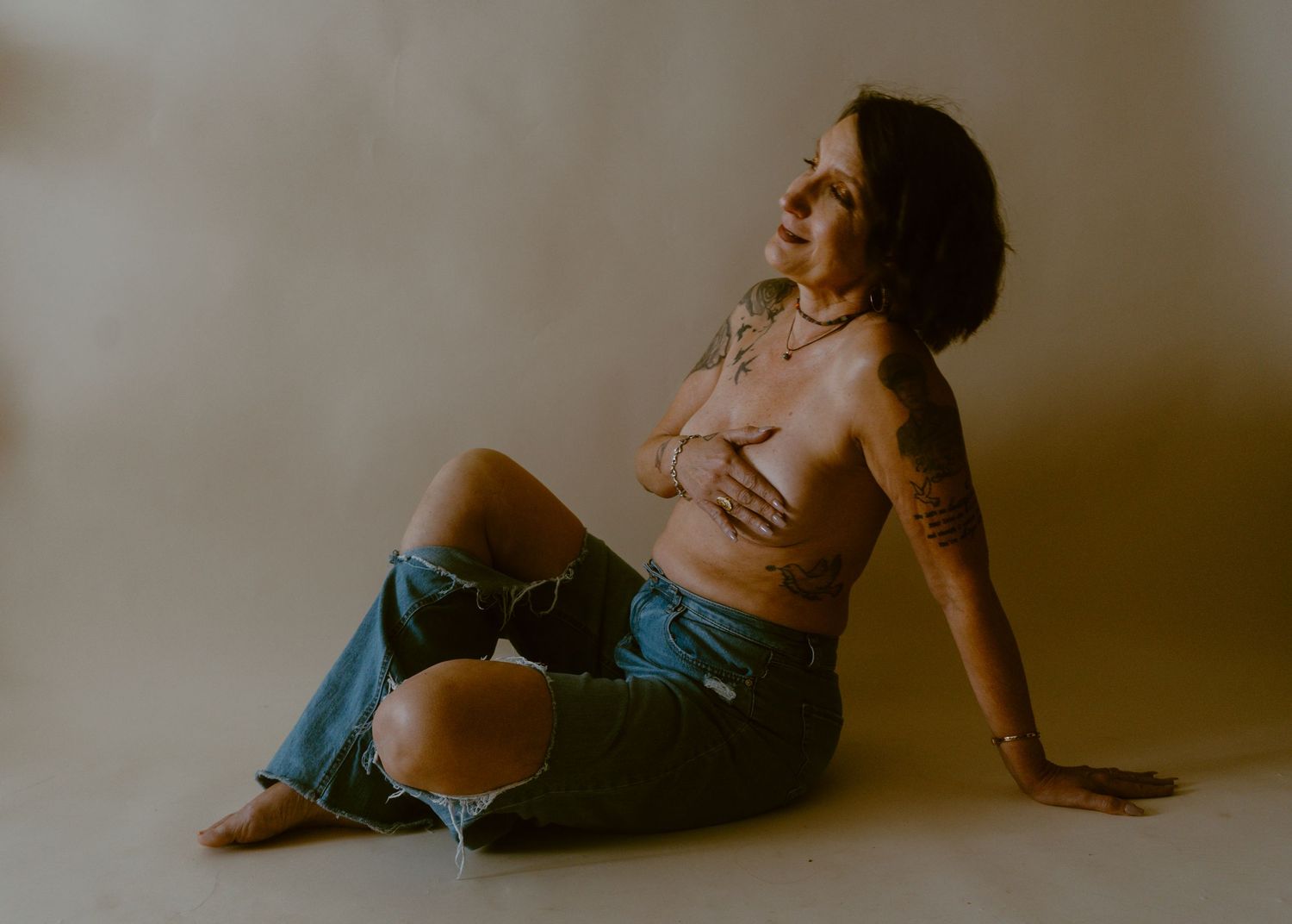 Woman with tattoos sitting on floor in denim skirt and crop top against neutral studio background.