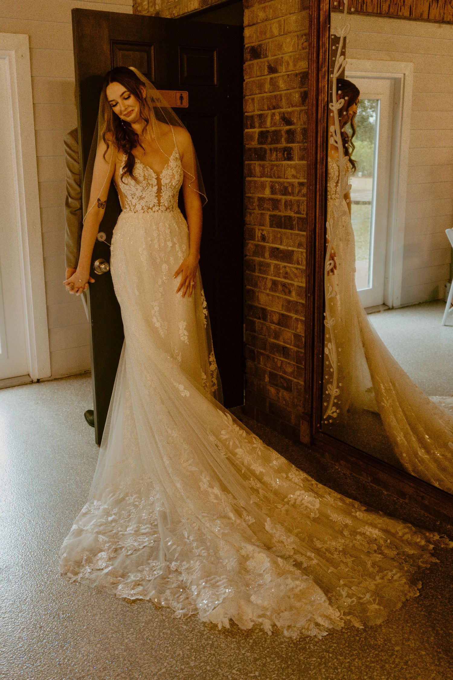 Bride in elegant lace mermaid wedding gown with long train standing by brick wall doorway.