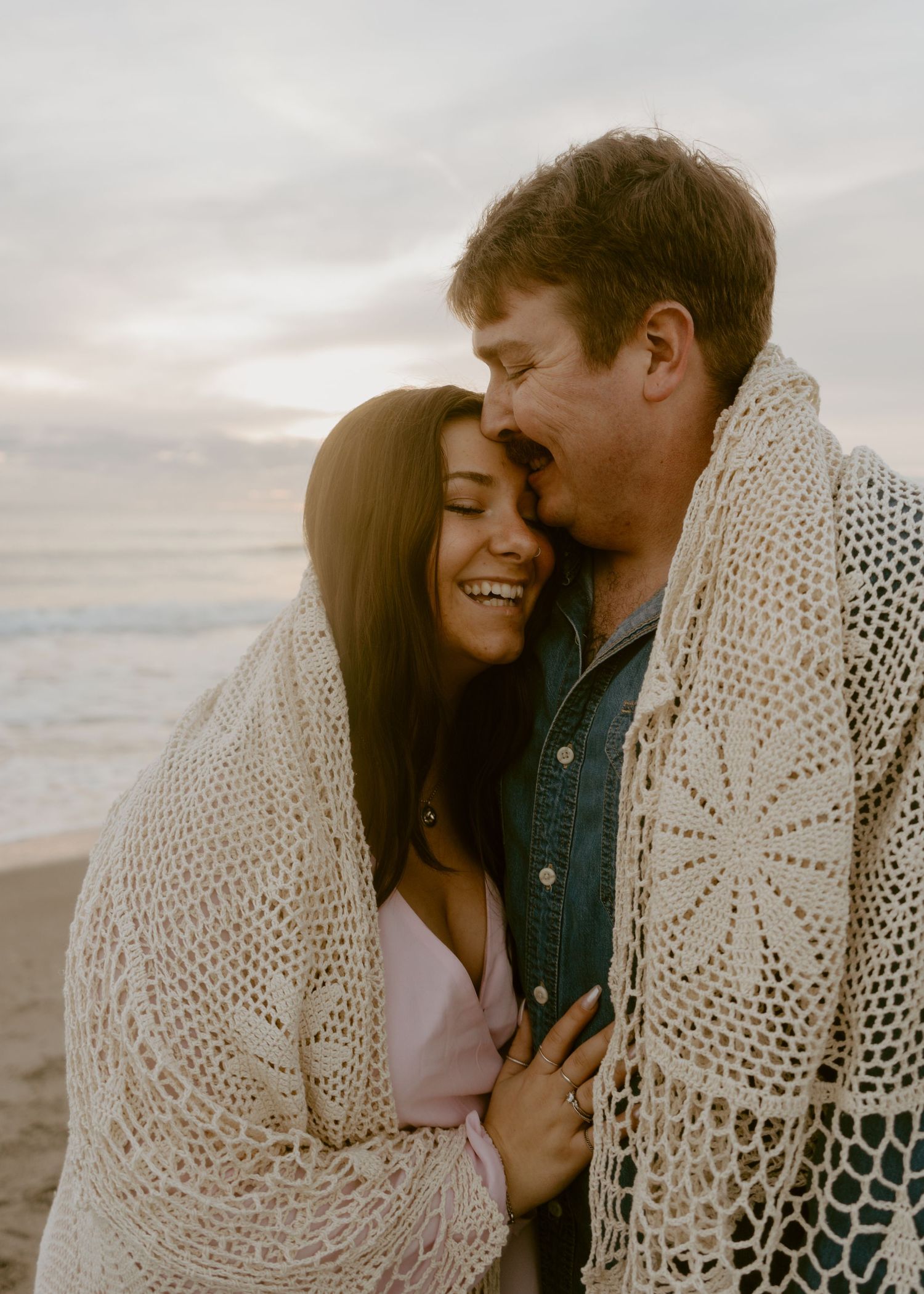 Couple wrapped in crochet blanket sharing a romantic moment on the beach at sunset.