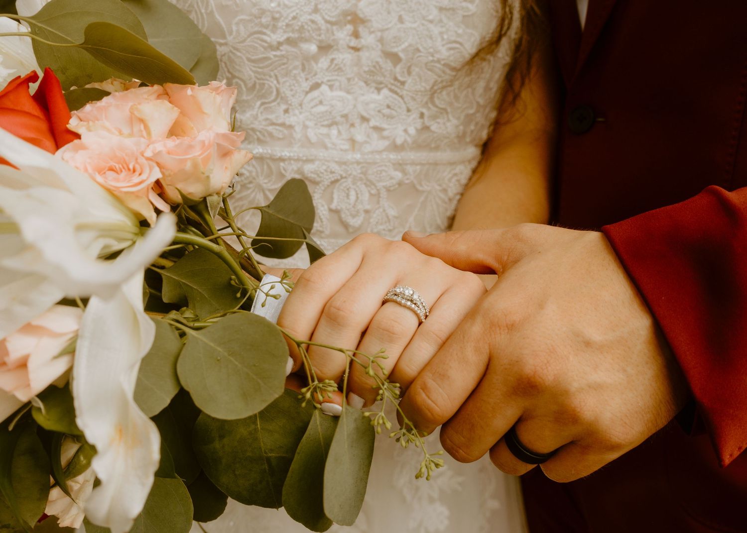 Bride and groom holding hands with wedding rings visible, bride holding a bouquet of pink roses and eucalyptus.