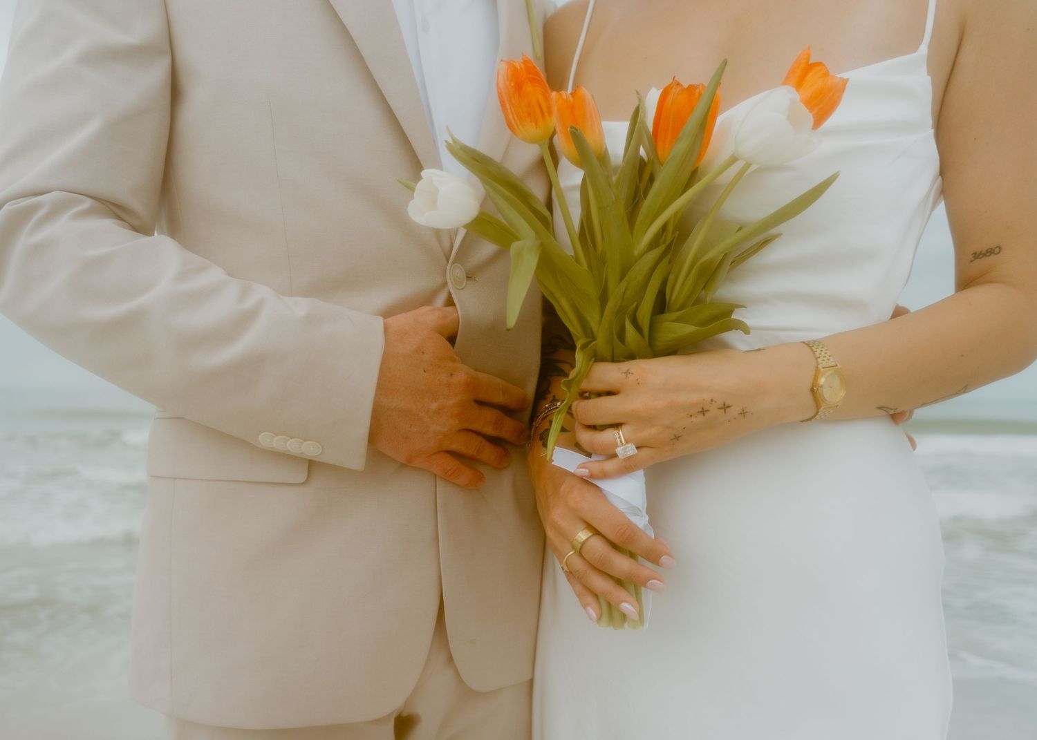 Couple holding orange and white tulip bouquet in wedding attire on beach, bride in white dress groom in beige suit.